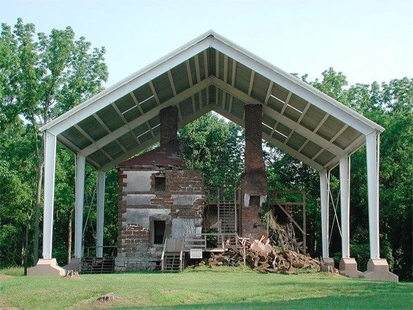 Restoration of a historic brick building with two brick chimneys under a modern white-roofed shelter in a park.