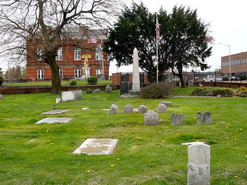 A historic graveyard with small headstones, a white obelisk monument, and a statue of a woman in front of a red brick building with flags in the background.