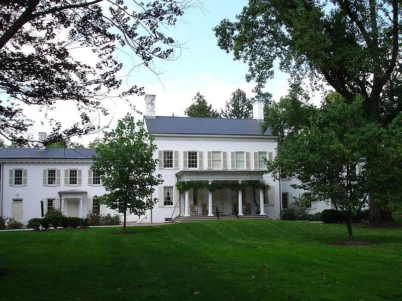 A large white historic house with multiple stories, surrounded by green trees and a well-maintained lawn.