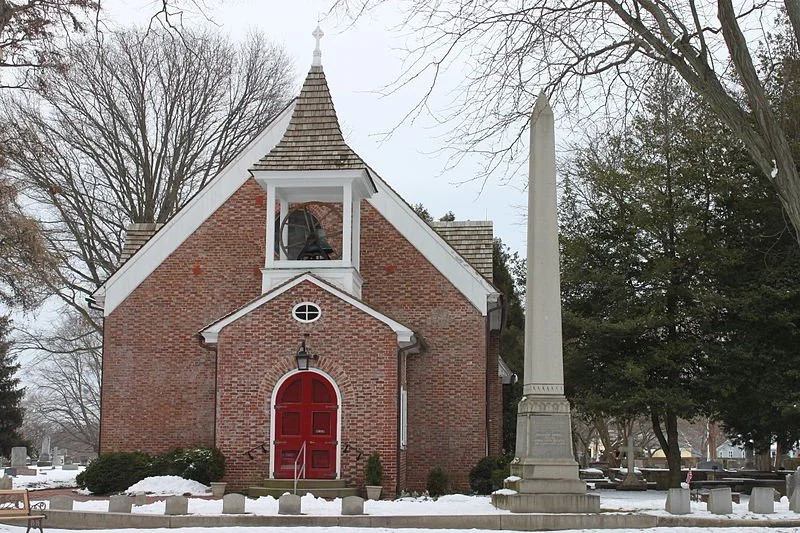 A brick church with a red door and a steeple, surrounded by snow and trees.