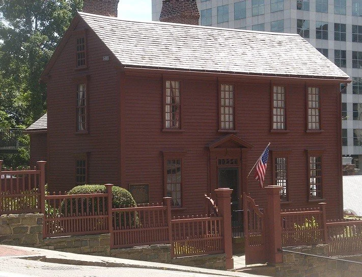A historic, two-story, red wooden house with a gabled roof, surrounded by a matching red fence with a gate. An American flag is mounted near the entrance, and modern buildings are visible in the background.