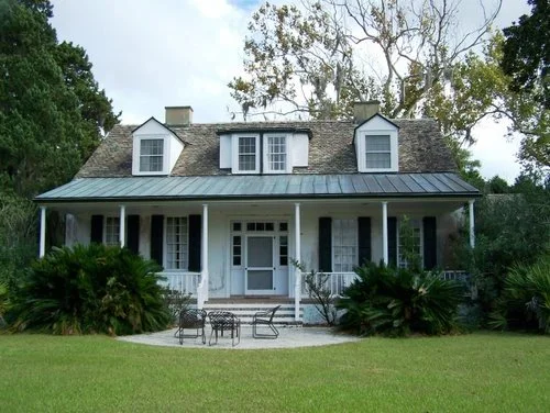 Front view of a two-story house with a covered porch, white exterior, and dormer windows, surrounded by a lawn and trees.