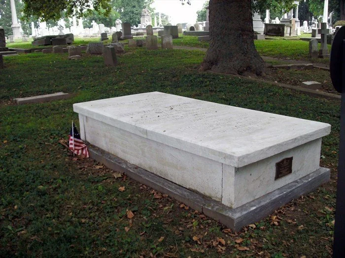 A white marble gravestone in a cemetery with an American flag at its corner and several other headstones in the background.