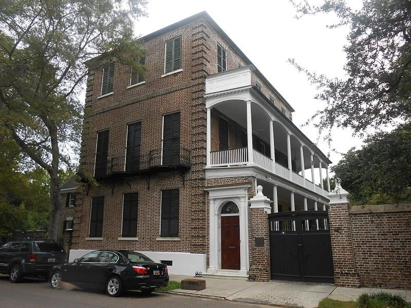 Three-story historic brick house with a large balcony on the second floor, white decorative columns, and a wooden front door. There are parked cars in front, and trees surrounding the house.