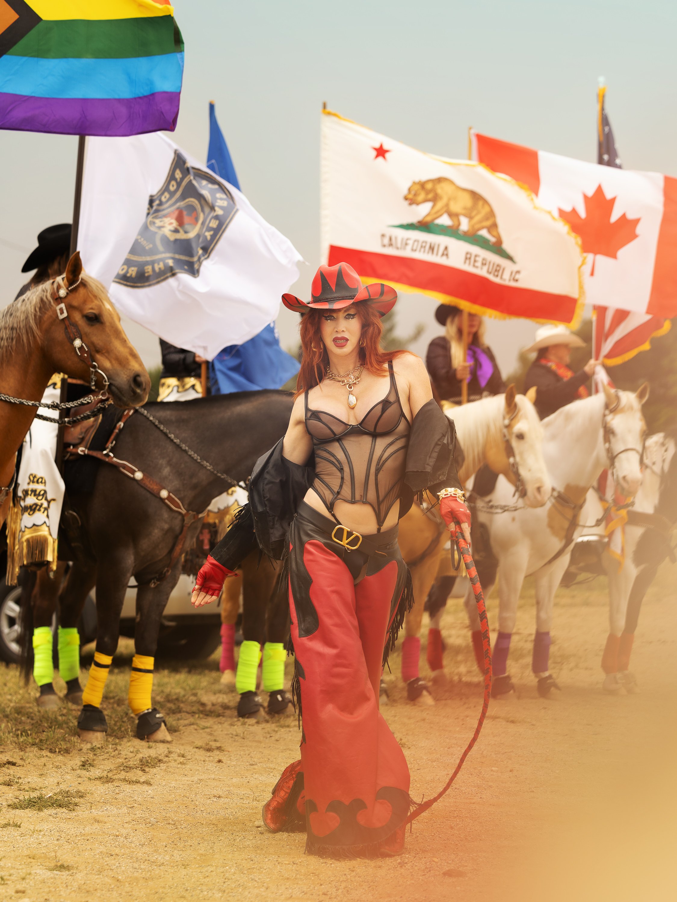 A woman dressed as a cowgirl with a black and red outfit and cowboy hat, walking in front of a line of horses and people holding flags for LGBTQ+ pride, California, and Canada.