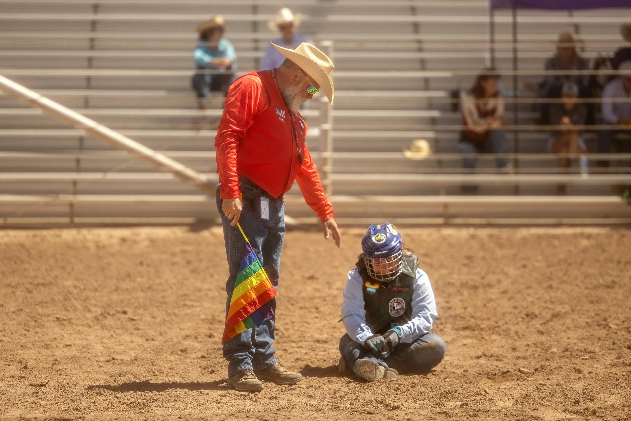 A cowboy, wearing a red jacket, cowboy hat, and sunglasses, is holding a small rainbow flag, standing next to a young person sitting on the dirt ground with a helmet and protective gear. The setting appears to be an outdoor arena with bleachers and s