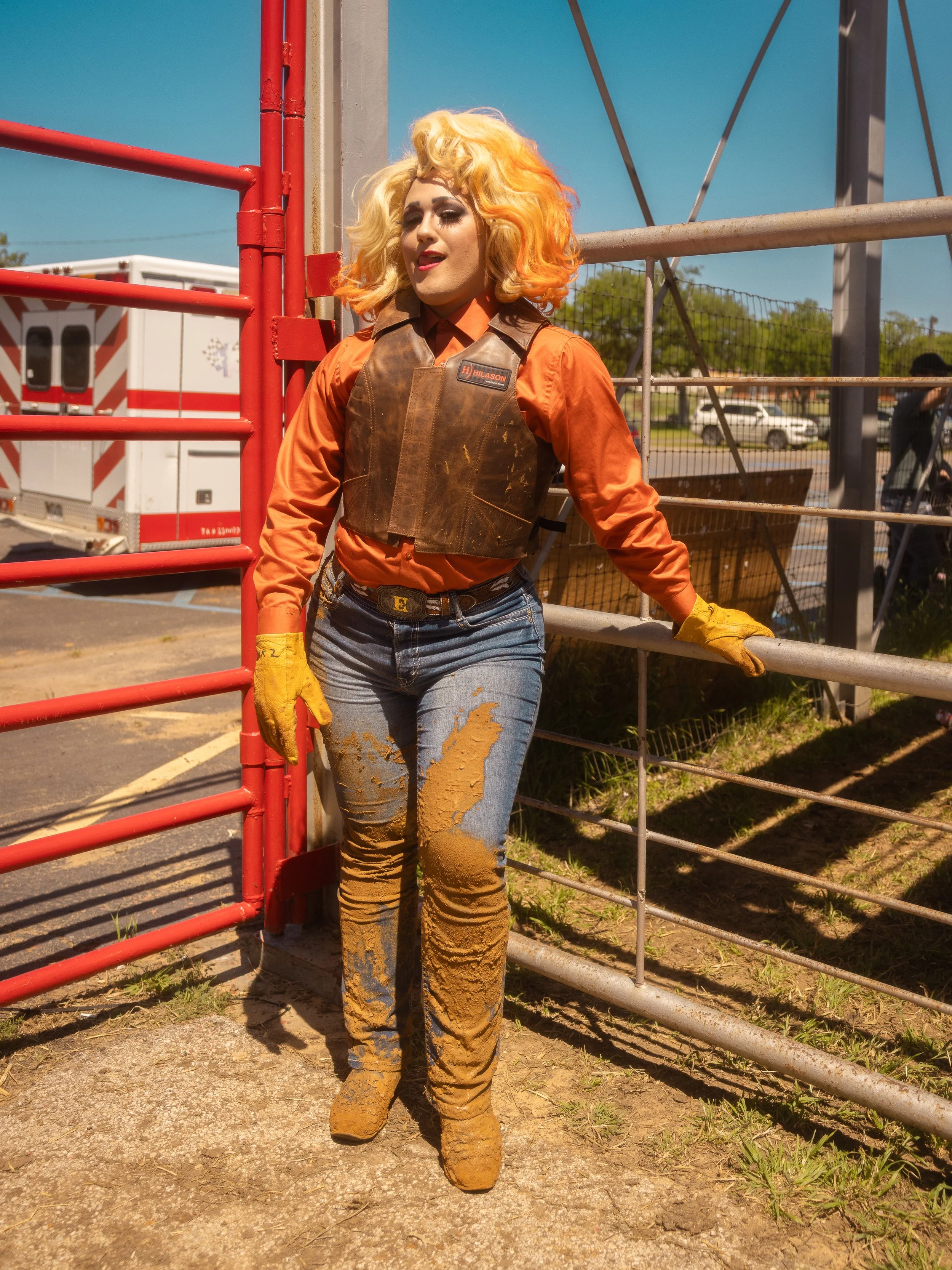 A woman with curly, blonde hair and orange-colored hair highlights, dressed in a dusty orange shirt, brown vest, and jeans, standing outdoors near a red and beige metal fence, with mud on her pants and boots, during daytime.