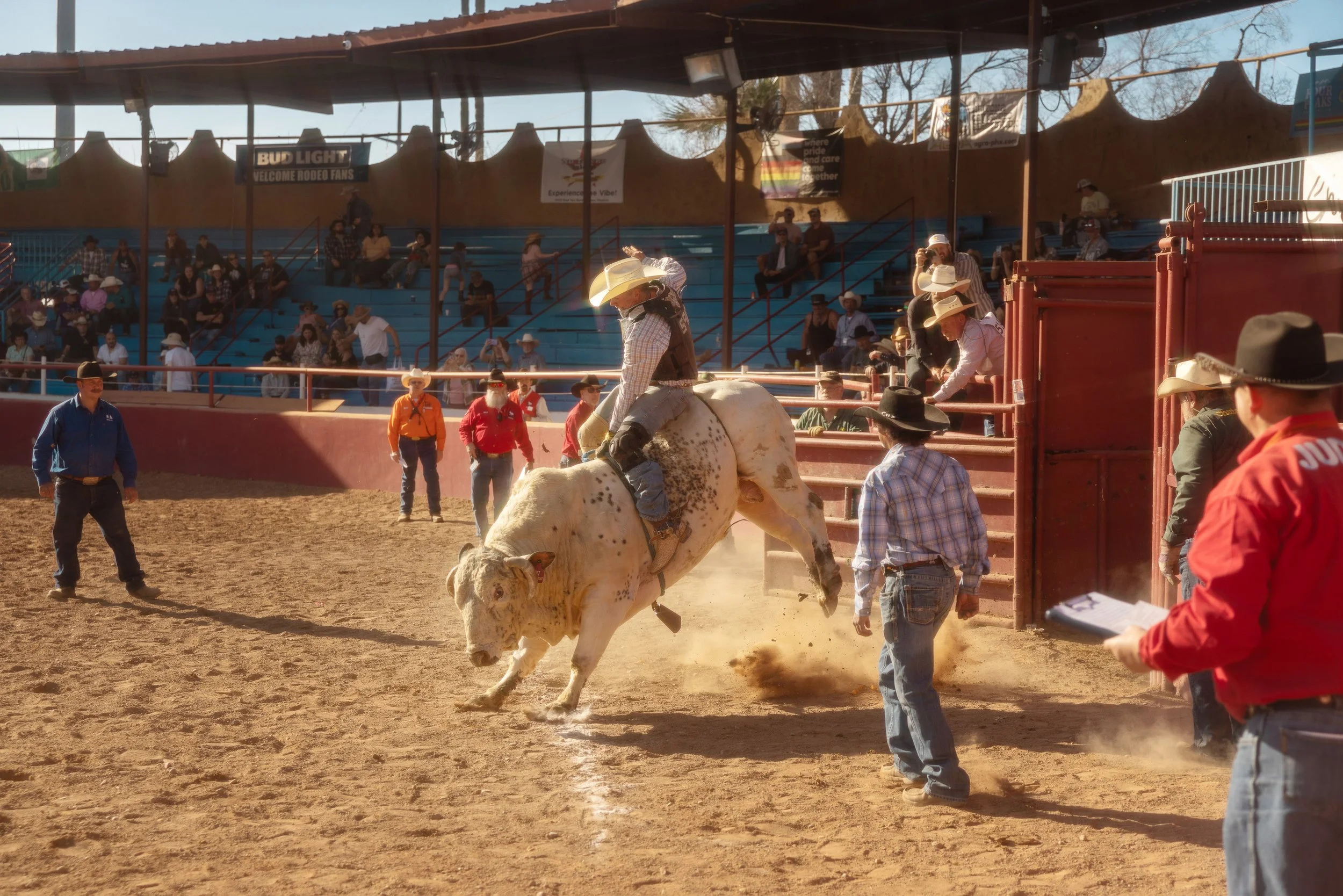 A rodeo scene with a cowboy riding a bucking bull, surrounded by spectators, officials, and other cowboys in western attire at an outdoor arena.