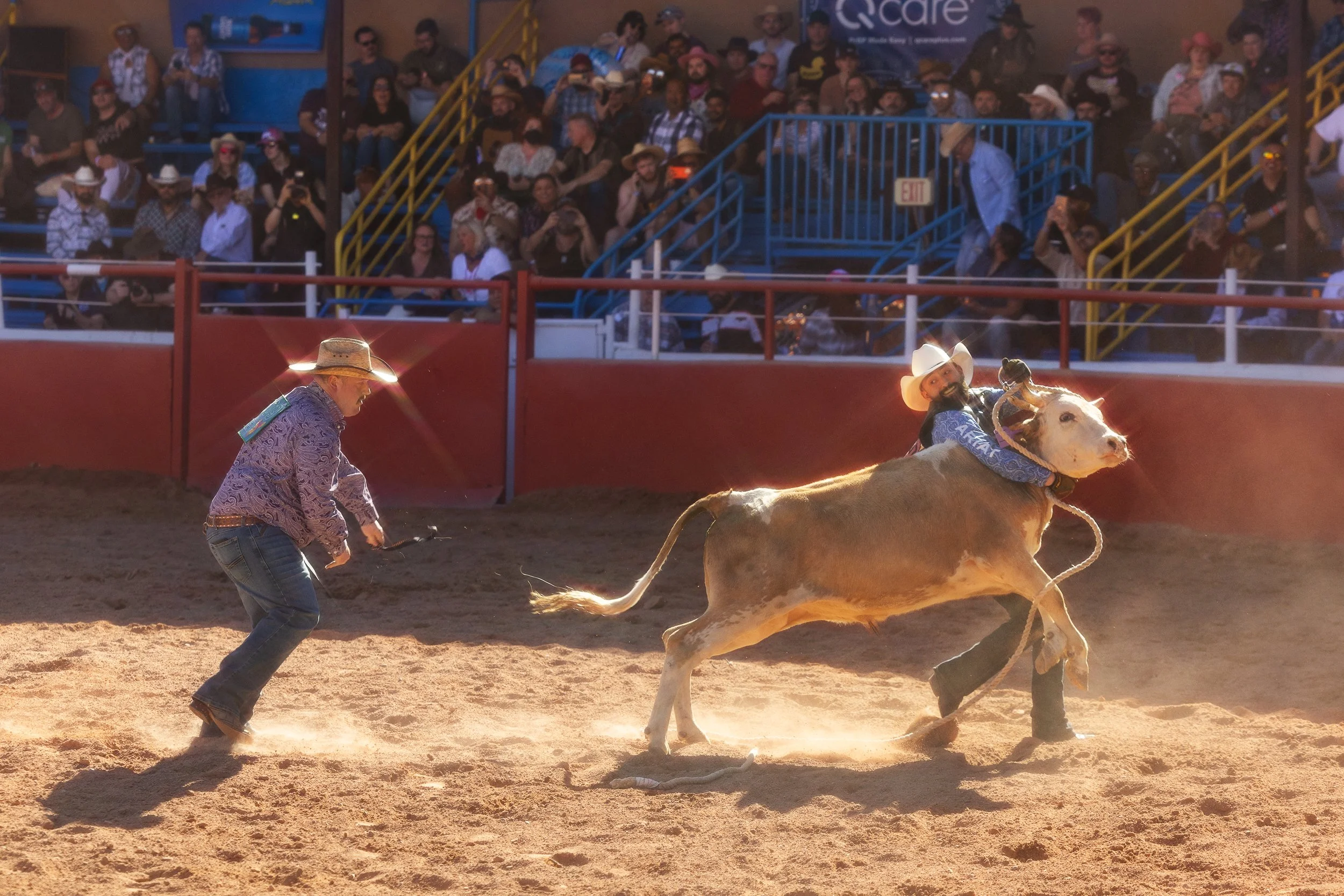 A rodeo arena with a man holding a lasso and a bull with a cowboy on its back. Spectators watch from the stands.