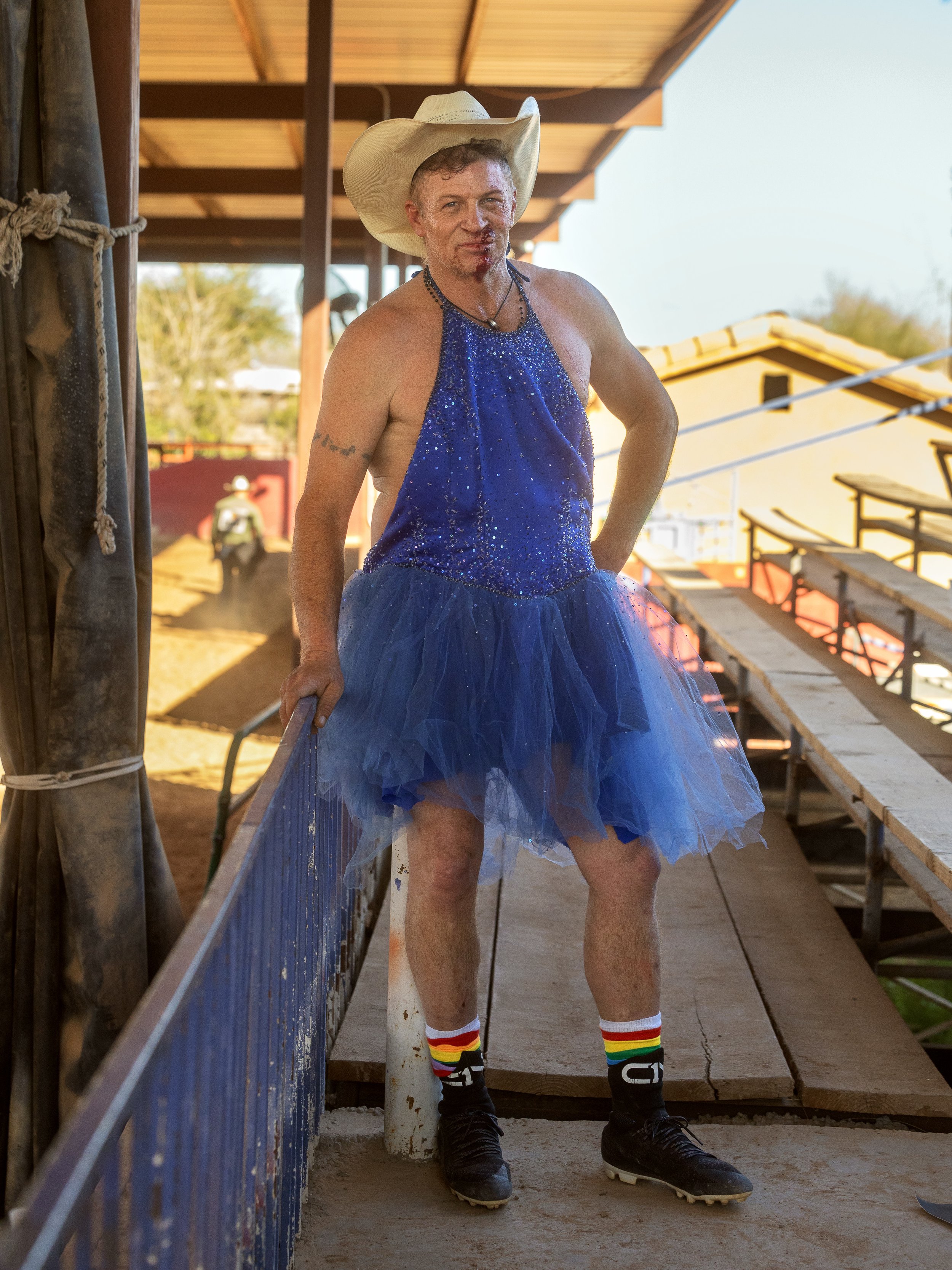 A man dressed in a blue sequined tank top, blue tutu, rainbow socks, and cricket shoes, wearing a cowboy hat, standing on a sports arena walkway.
