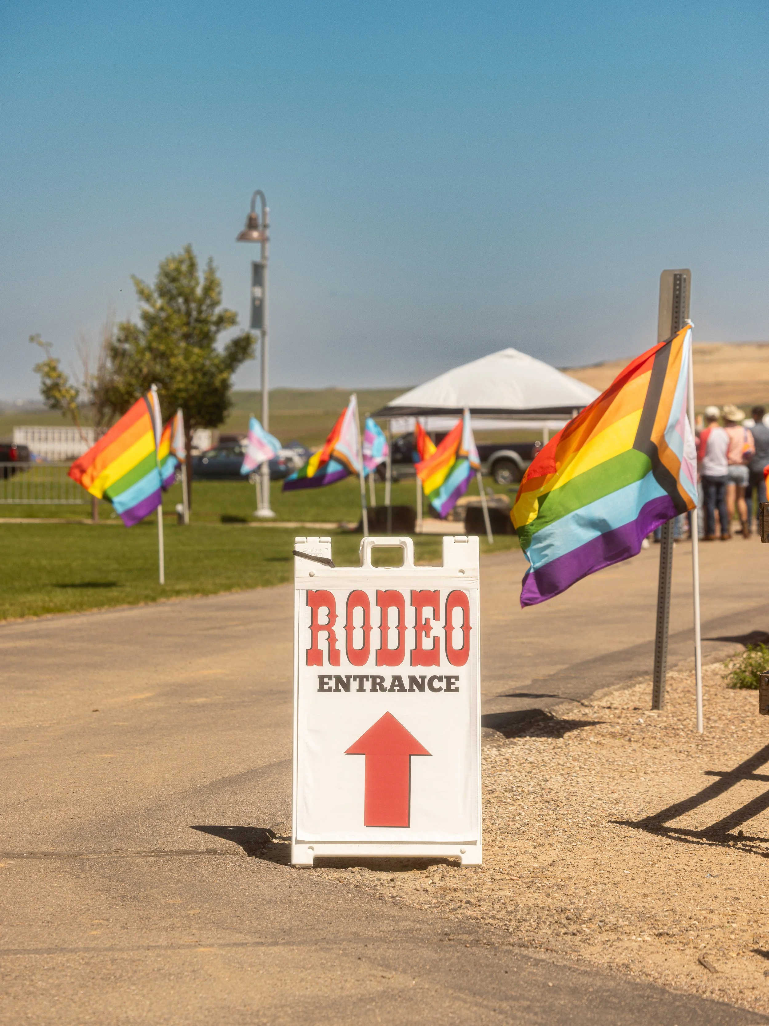 Sign for rodeo entrance with an arrow pointing upward, surrounded by rainbow flags, under a clear sky.