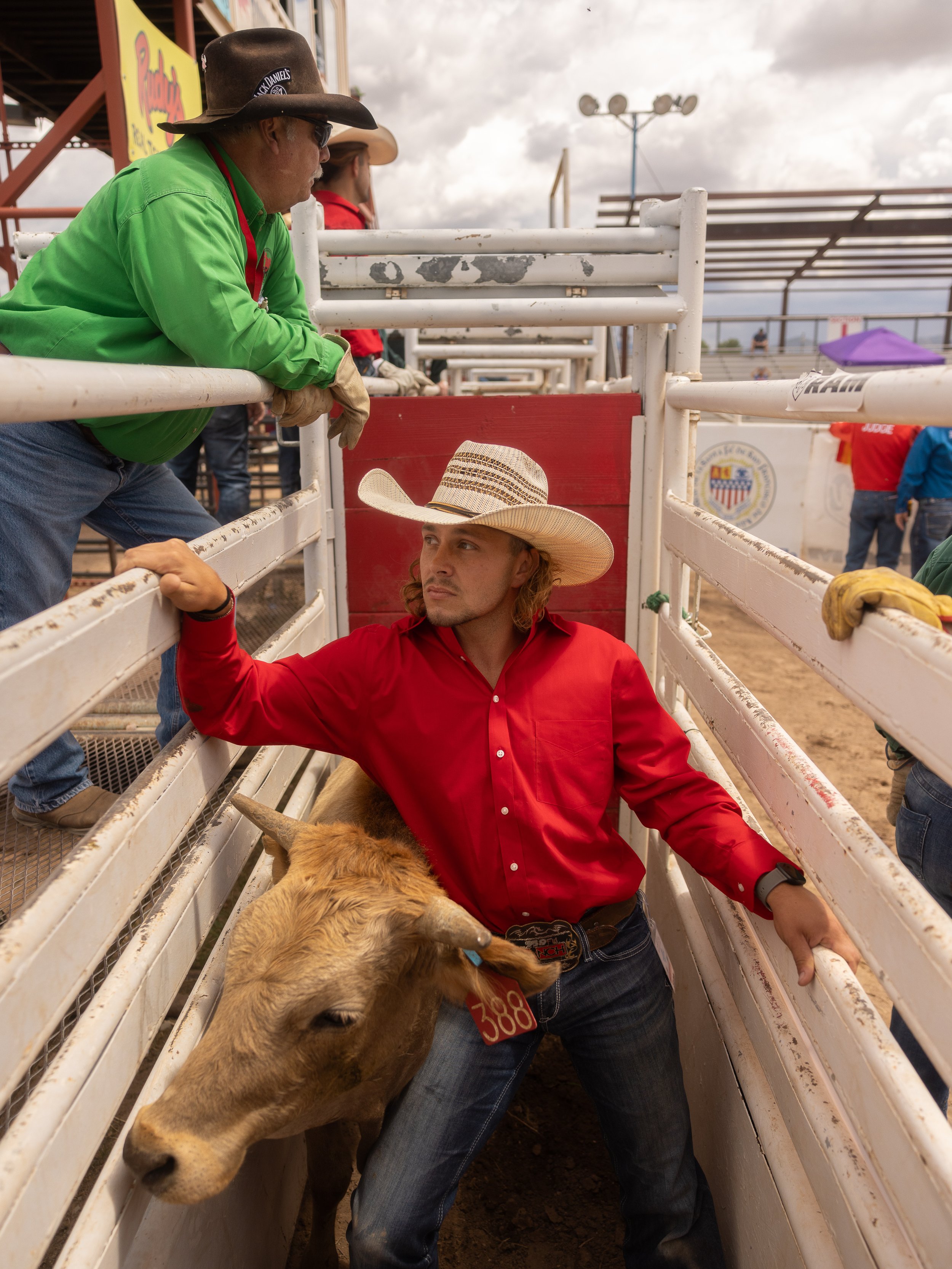 A young man in a red shirt and cowboy hat standing in a cattle pen with a calf, talking to a man in a green shirt and cowboy hat at a rodeo event.