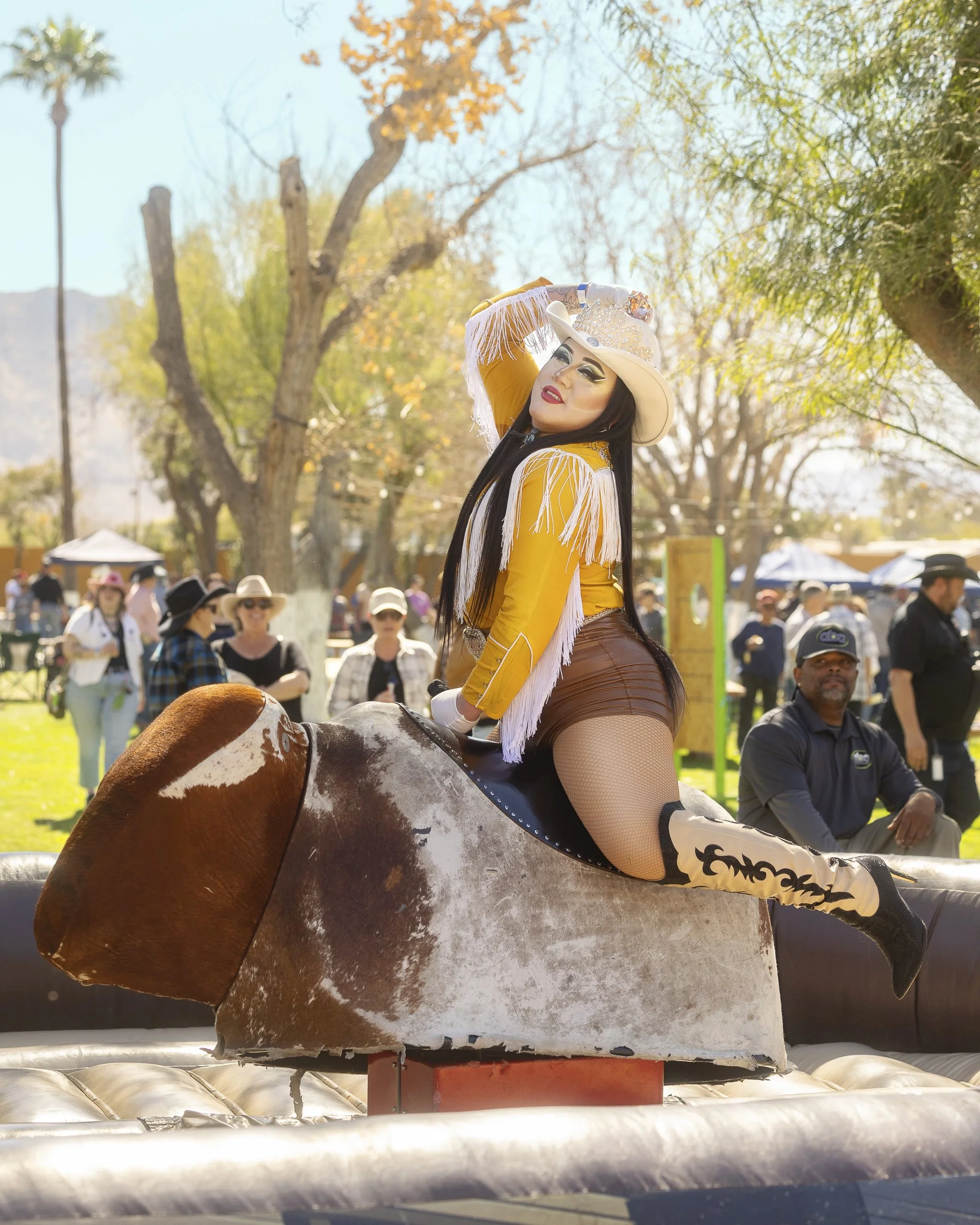 A woman in a cowboy costume posing on a mechanical bull at an outdoor fair or festival.
