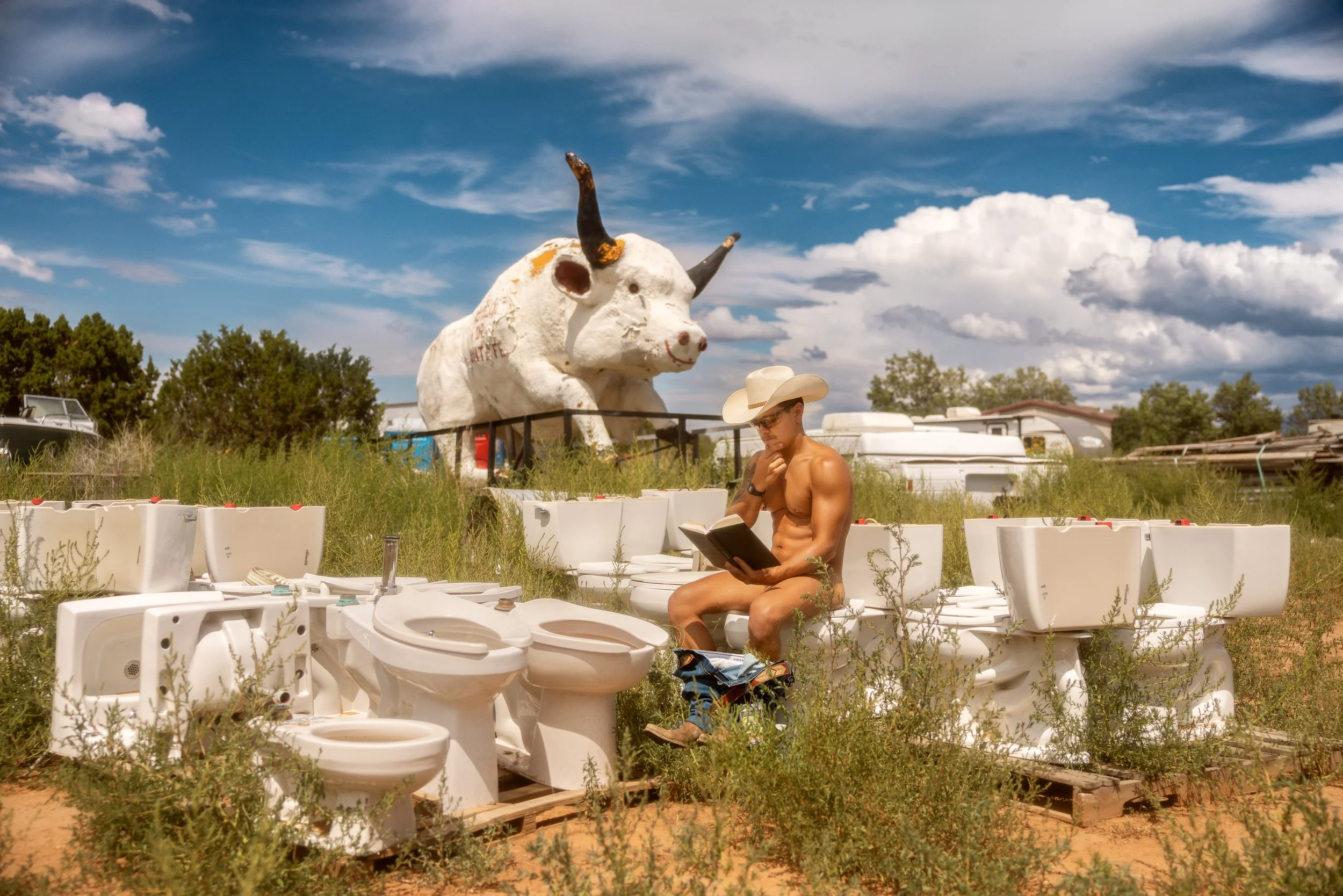 A shirtless man wearing a cowboy hat looking at a notebook while sitting amidst a display of toilets and bathroom fixtures in an outdoor lot with tall grass. In the background, there is a large cow sculpture with horns in front of a trailer and a blu