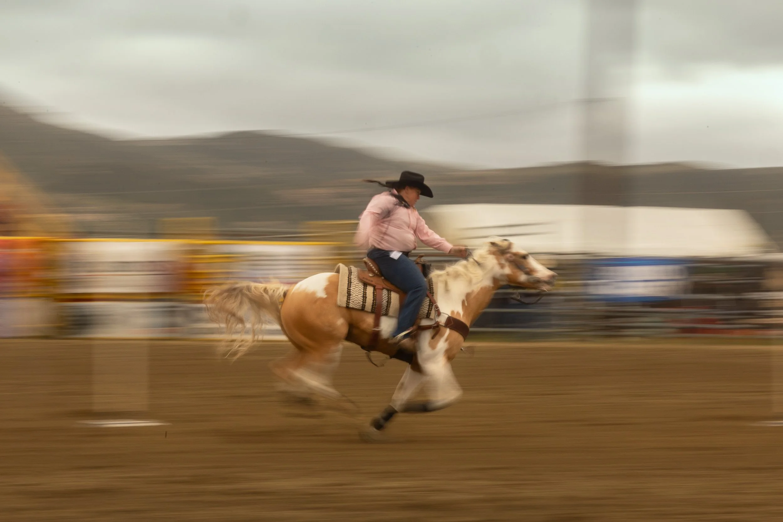 A woman wearing a cowboy hat and pink shirt riding a bucking horse in an arena with a mountain backdrop, with blurred motion indicating fast movement.