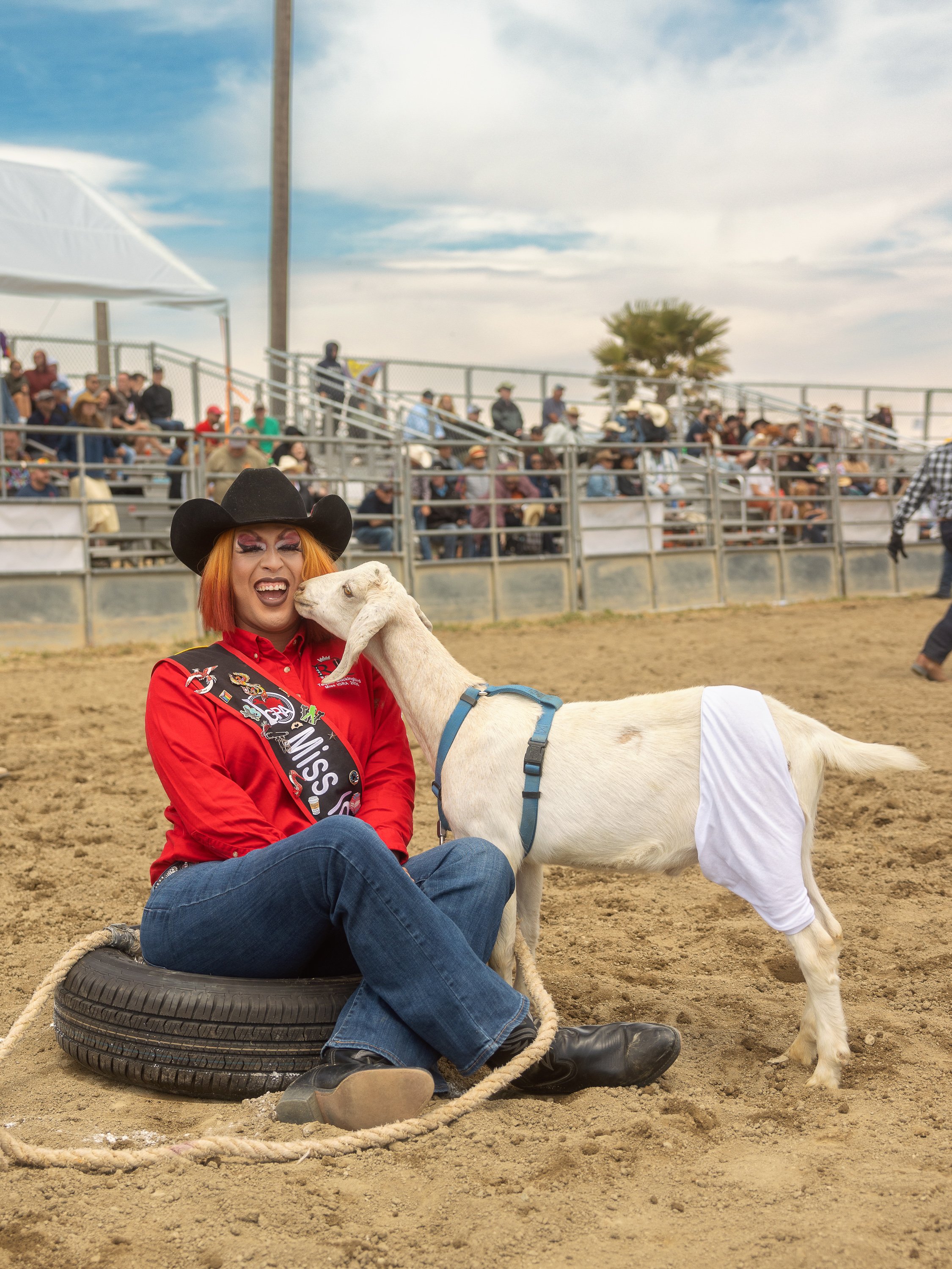 A person in a red shirt and black cowboy hat sitting on a tire, holding a goat that is nuzzling their face at an outdoor livestock event with a crowd in the background.