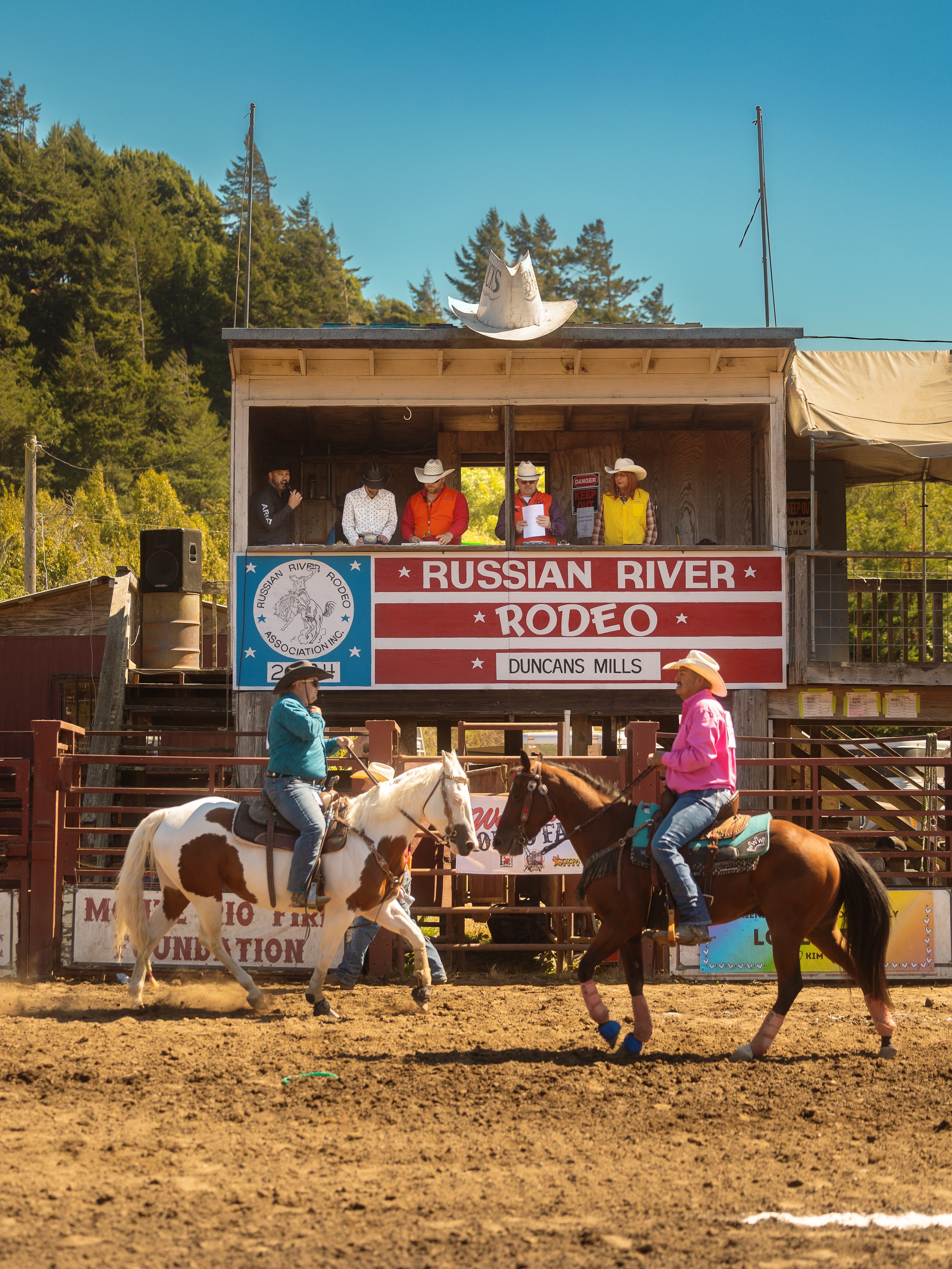 Two cowgirls riding horses in a rodeo arena with a wooden announcer's booth in the background, which has a large sign that reads 'Russian River Rodeo' and 'Duncans Mills'. The announcer's booth has four people wearing cowboy hats and western attire, 