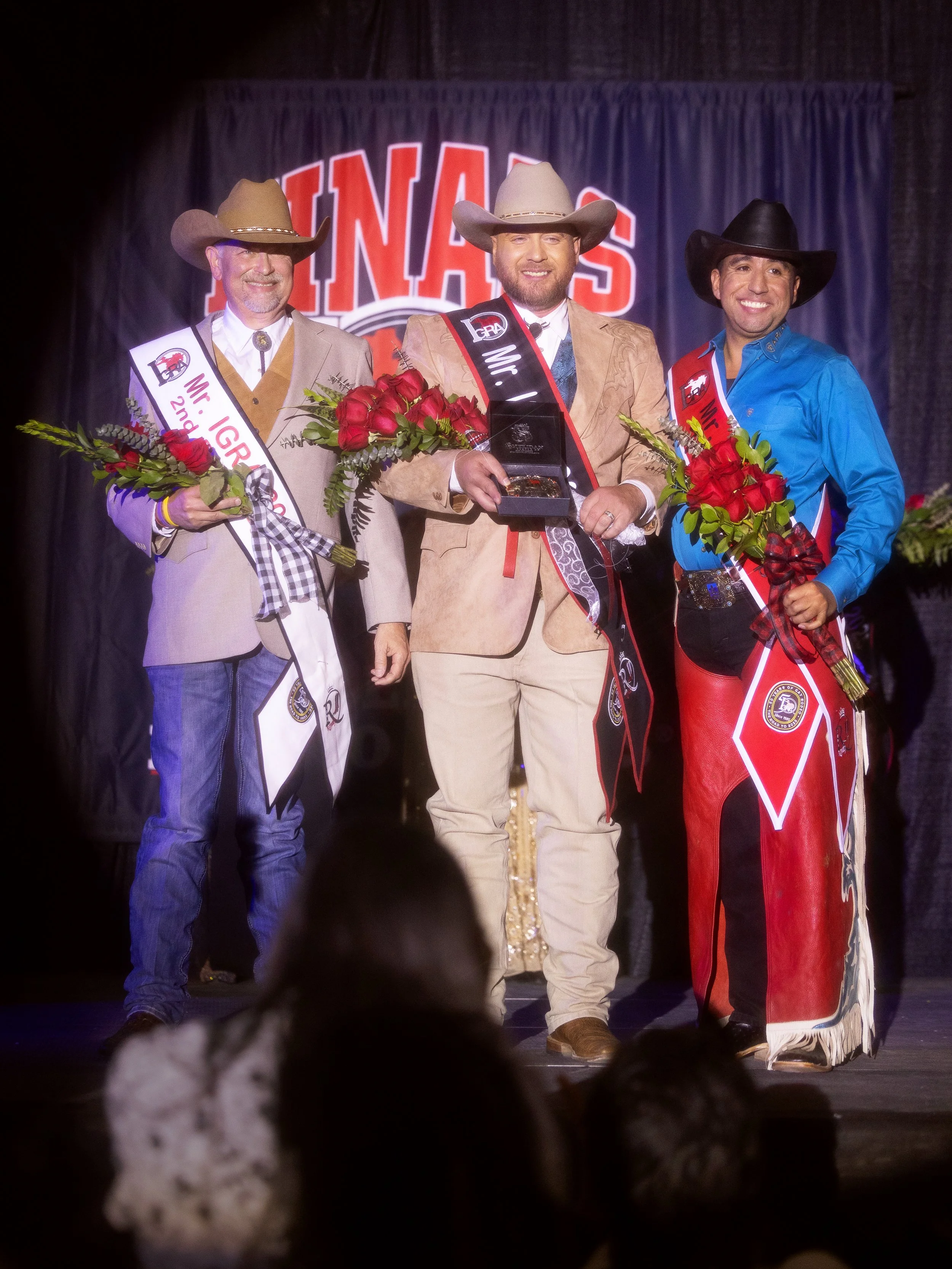 Three men dressed in western attire, standing on stage during a rodeo event, holding bouquets of flowers, with one man in the center receiving a trophy, all smiling and wearing cowboy hats and sashes indicating their titles.