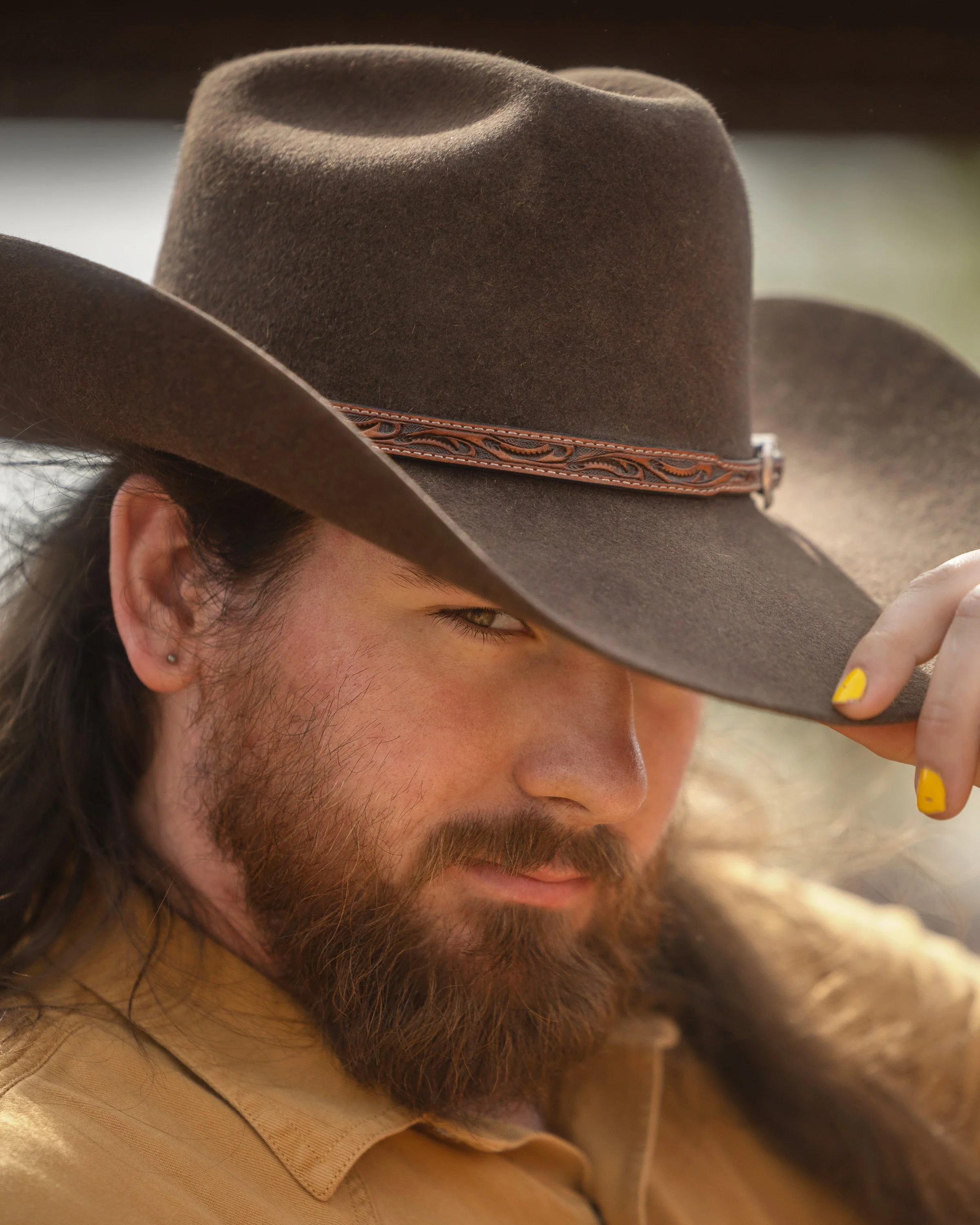 A man with a beard wearing a brown cowboy hat, looking downward, with yellow painted nails, and a tan shirt.
