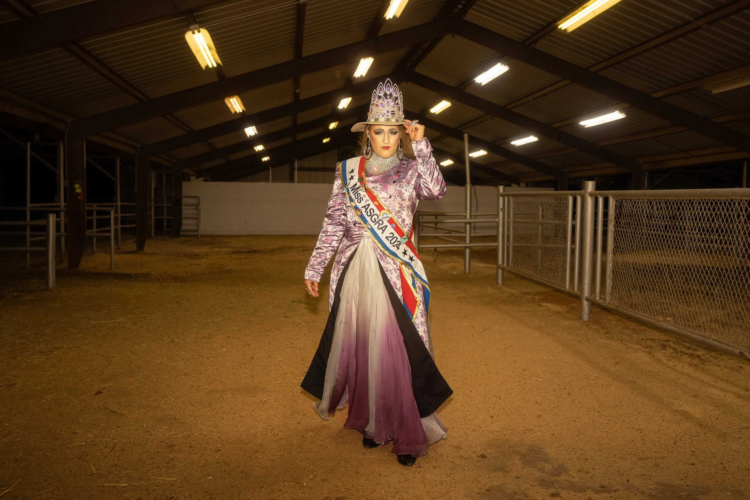 A woman dressed in an elaborate purple dress with a crown and sash reading 'Miss ASGRA 2024' standing inside a horse stable.