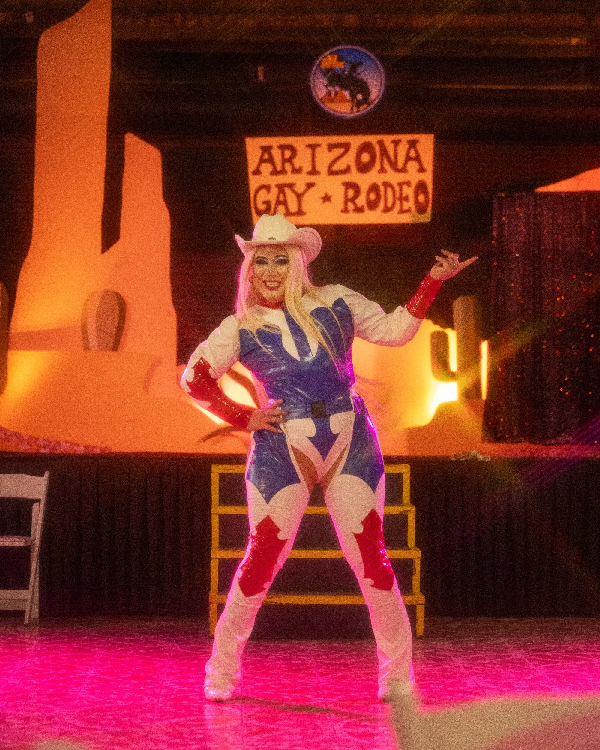 Performer dressed in a colorful outfit with a cowboy hat, standing on stage at the Arizona Gay Rodeo, with a backdrop sign reading 'Arizona Gay Rodeo' and a western-themed background.