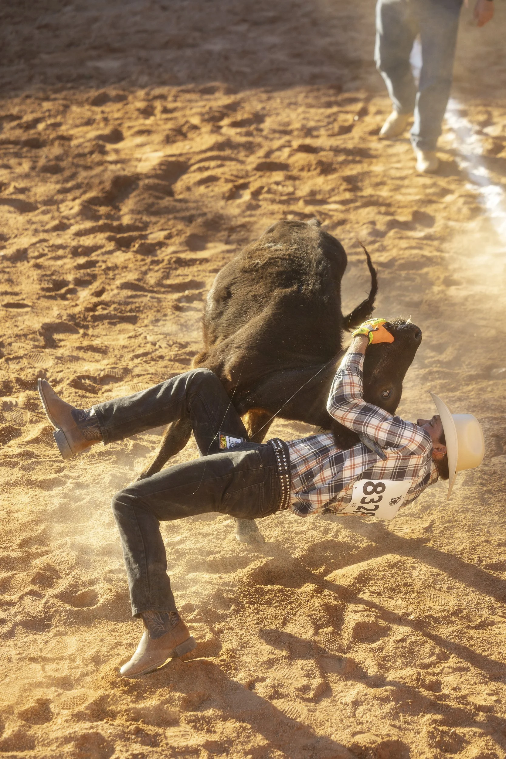 A person wearing western attire and a cowboy hat is falling off a bucking bull during a rodeo event on sandy ground, with other person and bull in the background.
