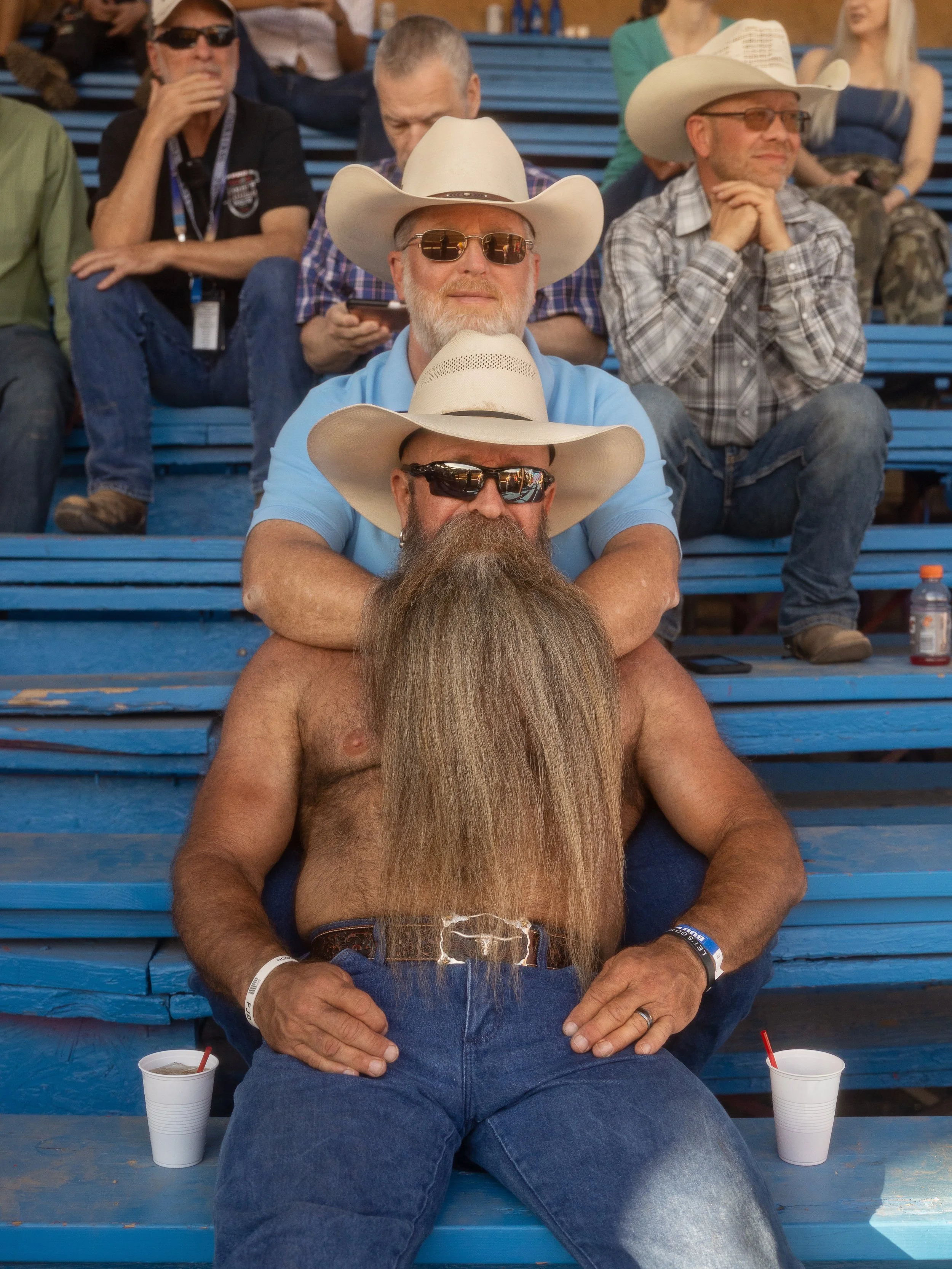 A shirtless man with a long beard, wearing sunglasses and a cowboy hat, sitting on blue bleachers at a sporting event with a group of people, some wearing cowboy hats and sunglasses.