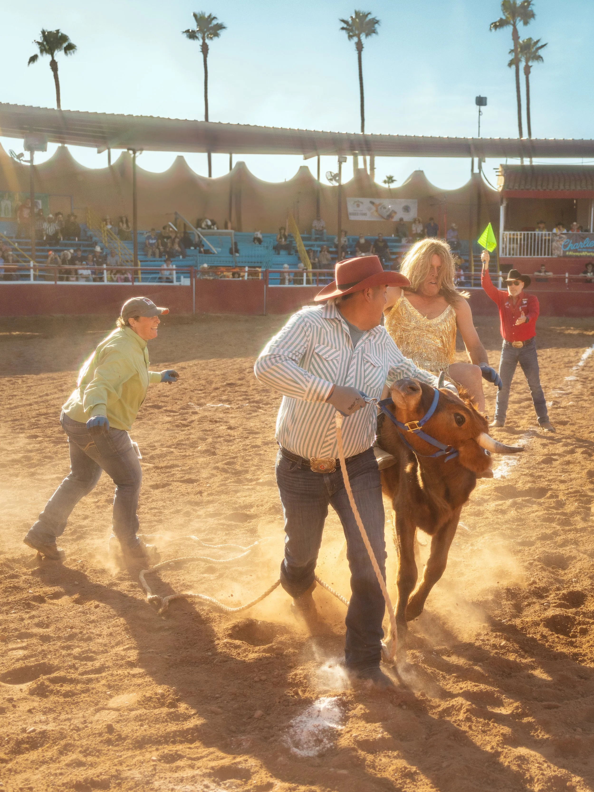 A rodeo scene with a cowboy on a bull or steer, two women, and a person holding a green flag, all on a dirt arena with spectators in the stands and palm trees in the background.