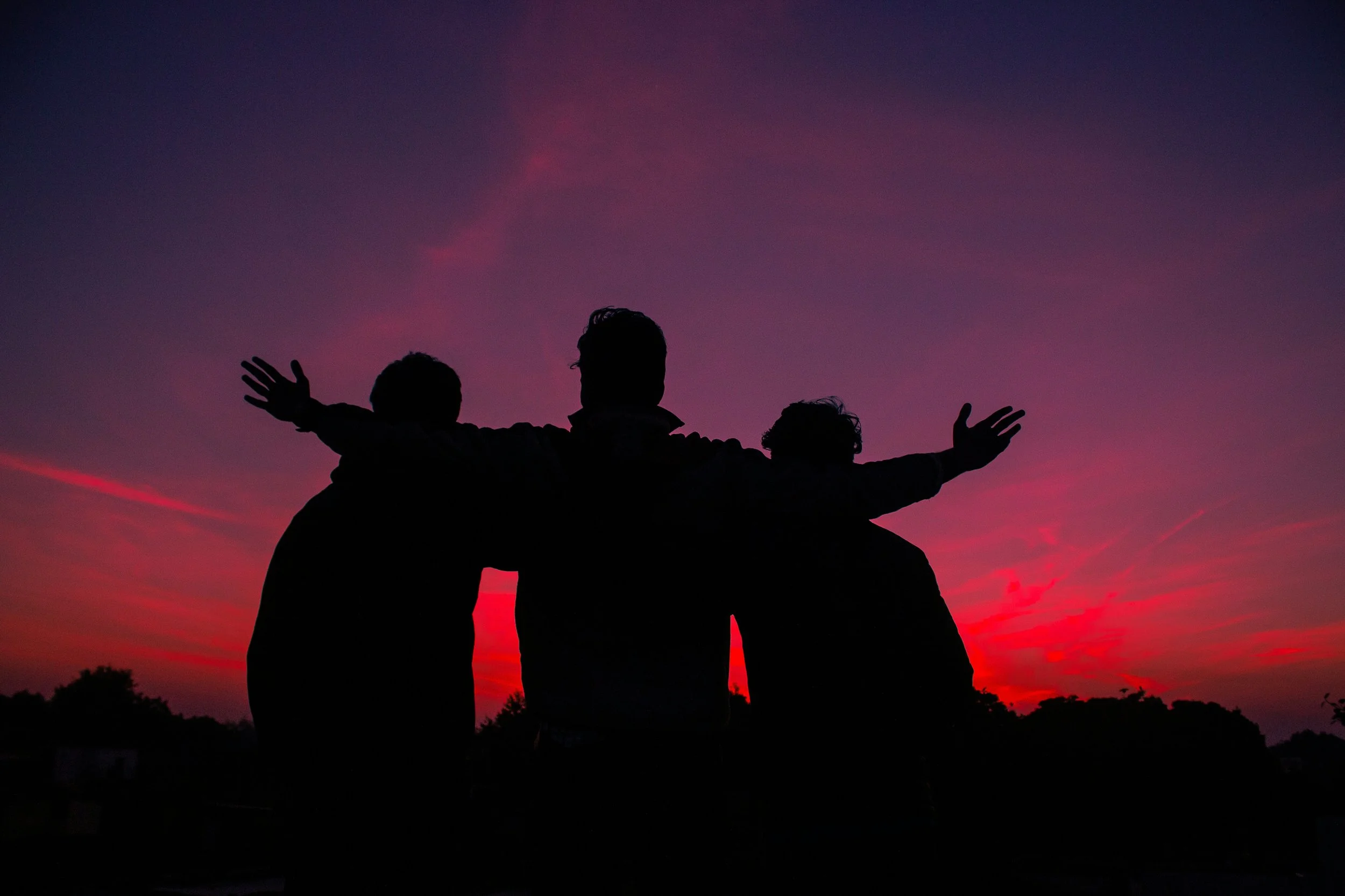 Silhouettes of three people standing together at sunset with their arms outstretched against a colorful sky with pink and purple hues.
