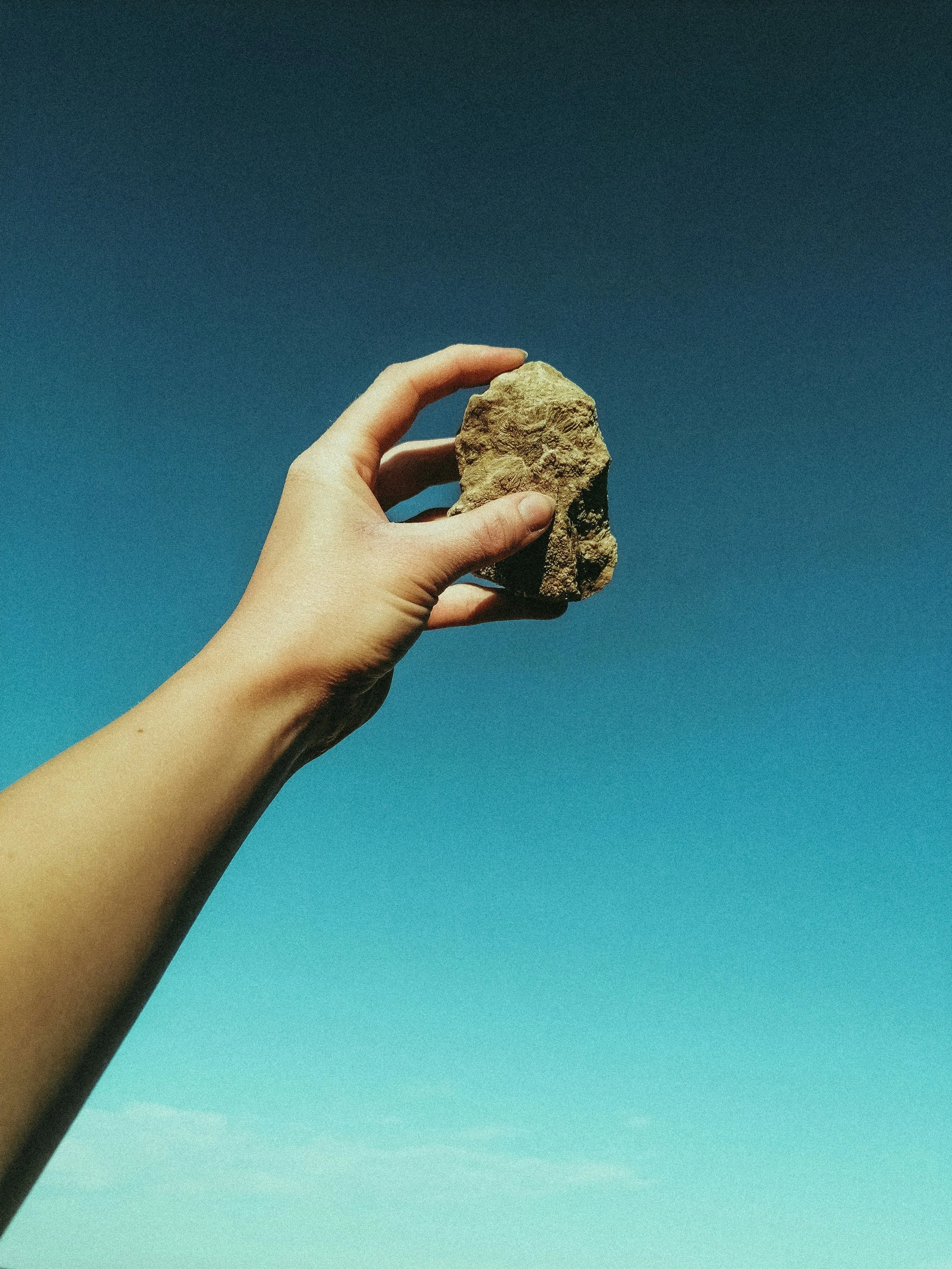 A hand holds up a rock against a clear blue sky.