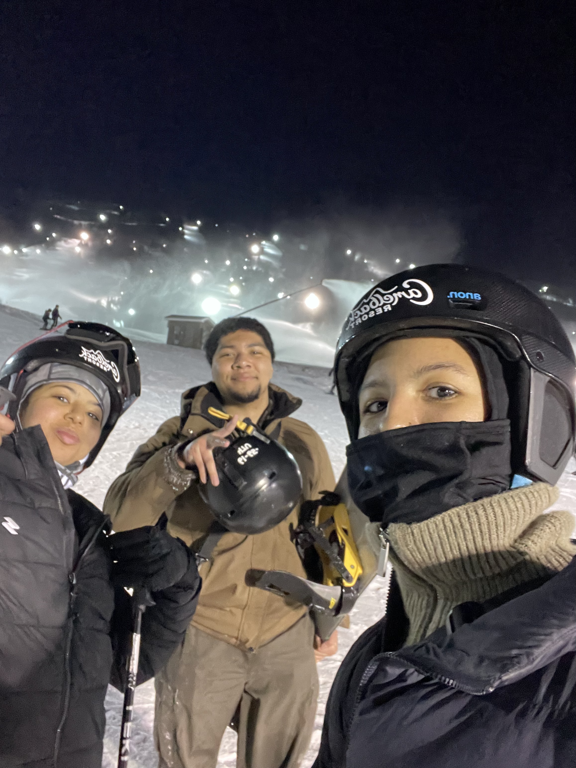 Three people in ski helmets and winter jackets on a snowy mountain at night, with ski slopes and bright lights in the background.