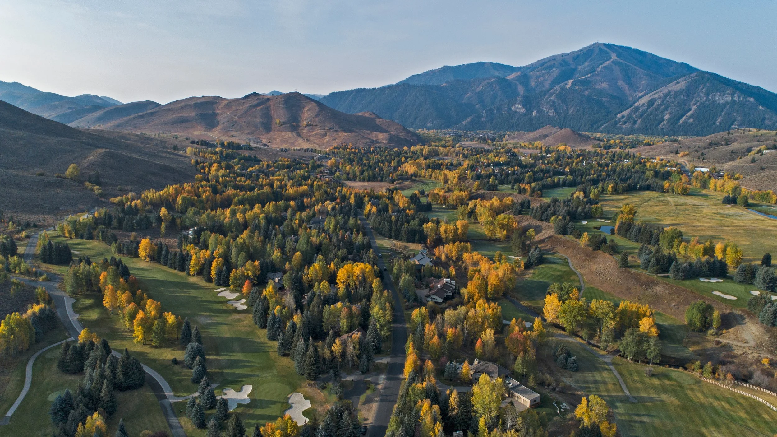 A landscape view of a lush green golf course with sand traps, surrounded by trees with fall foliage, mountains in the background, and a clear sky above.