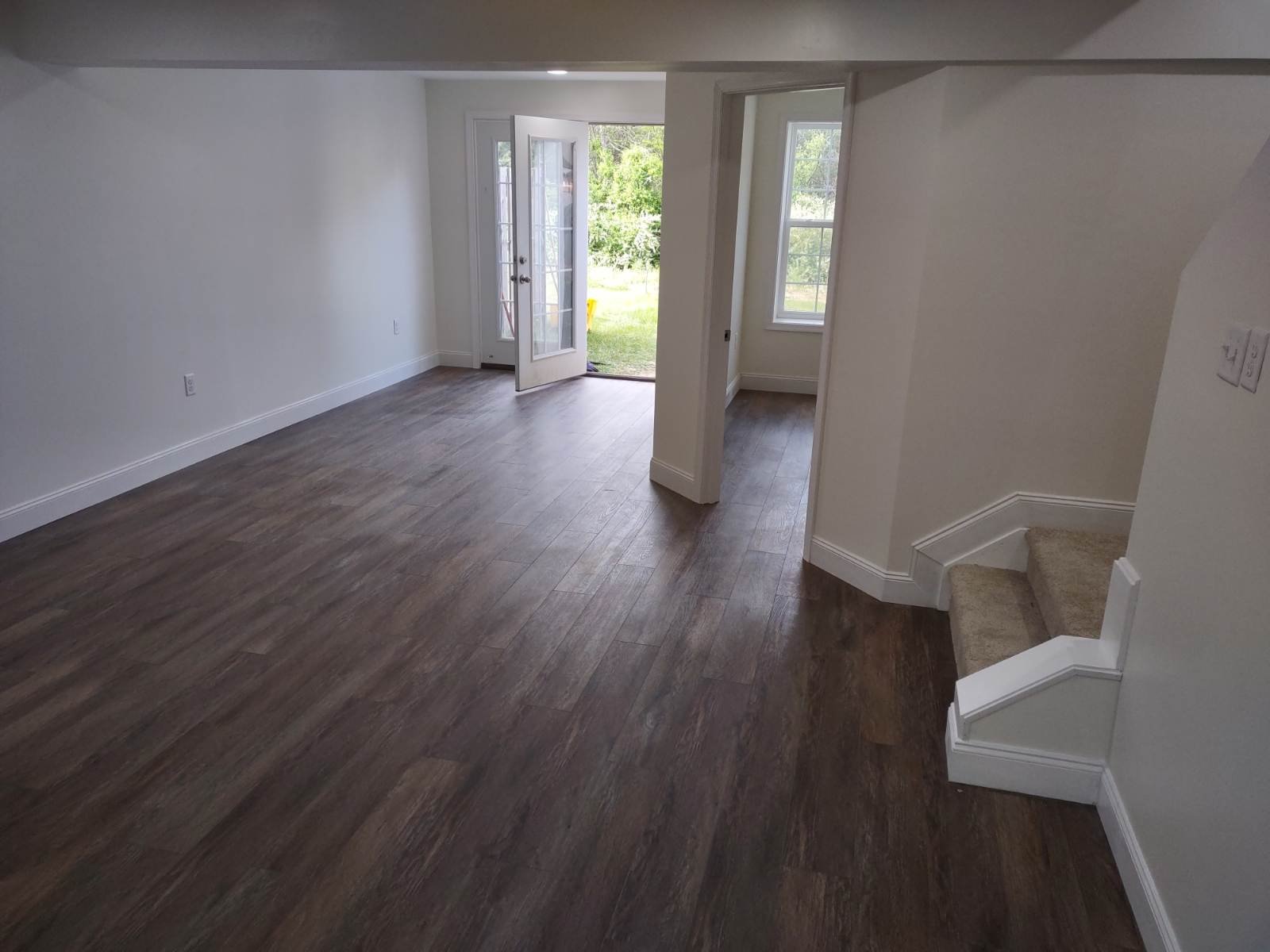 Empty living room with hardwood floors, white walls, a glass door leading outside, and a window near stairs with carpeted steps.