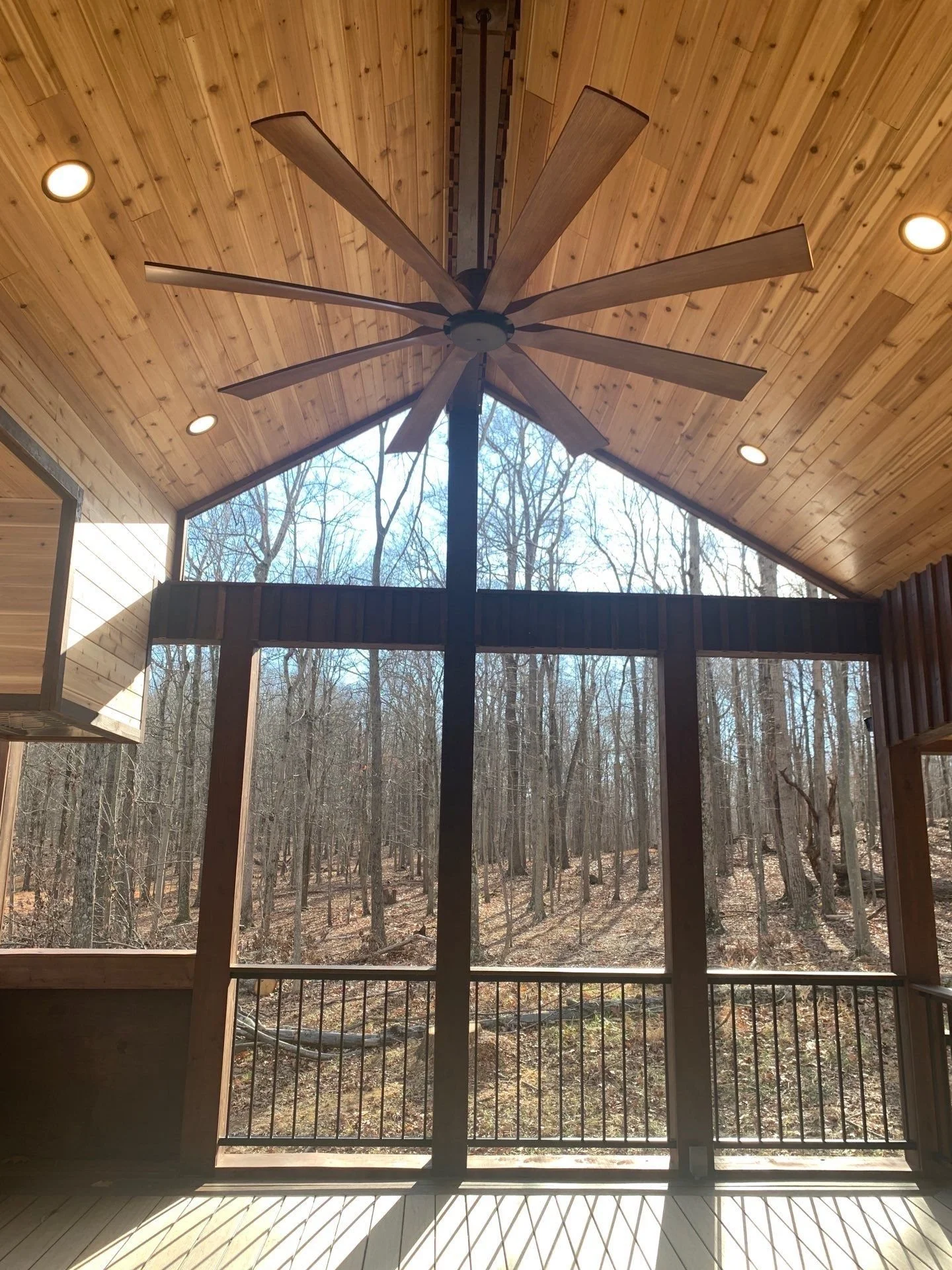 Interior view of a wooden porch or sunroom with a ceiling fan, recessed lighting, and large windows overlooking a wooded forest.