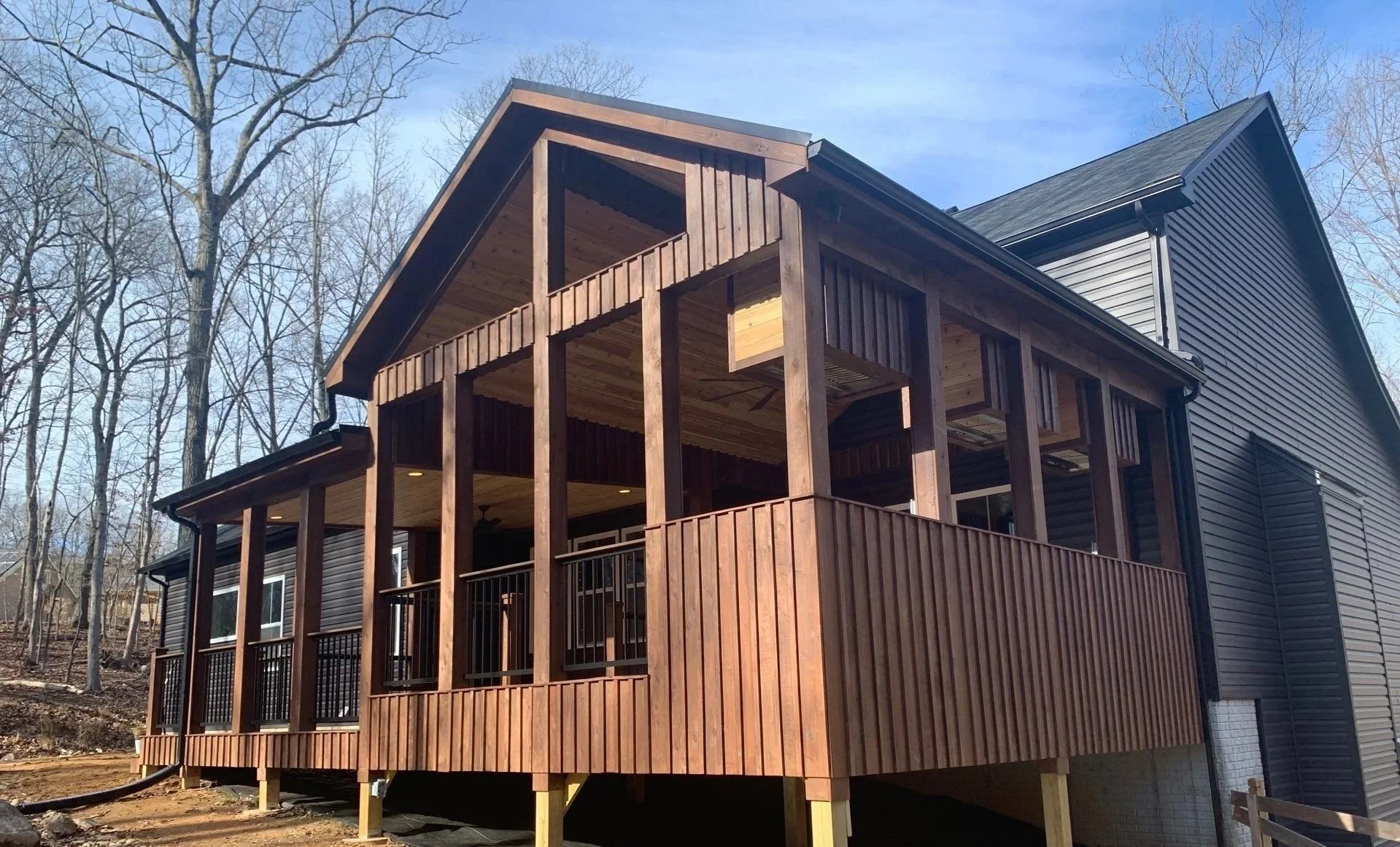 A two-story house with a large wooden porch on the upper level under construction, surrounded by trees with no leaves, and a partly cloudy sky.
