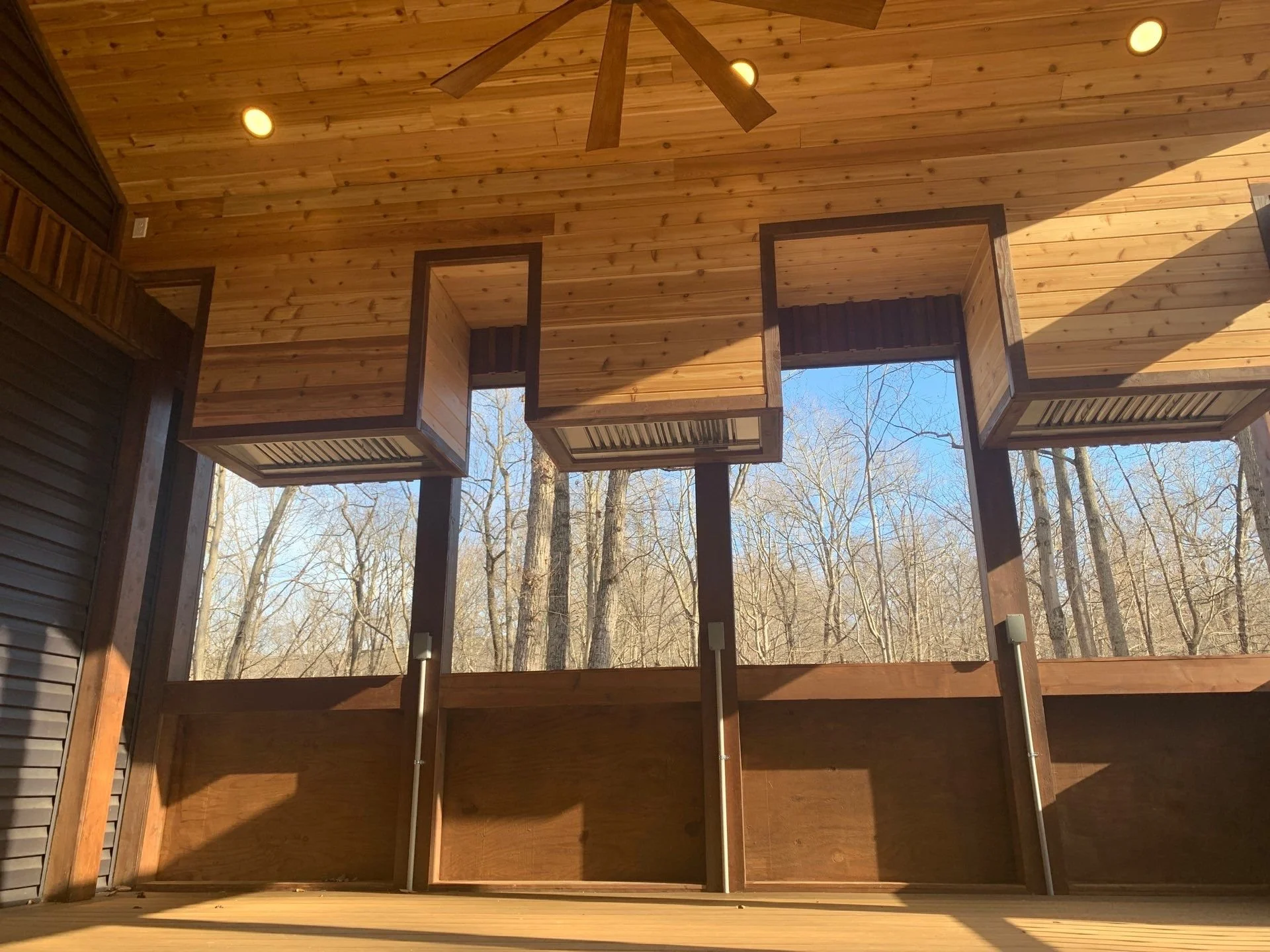 View of a wooden covered porch with open box-shaped compartments in the ceiling and a view of leafless trees outside.