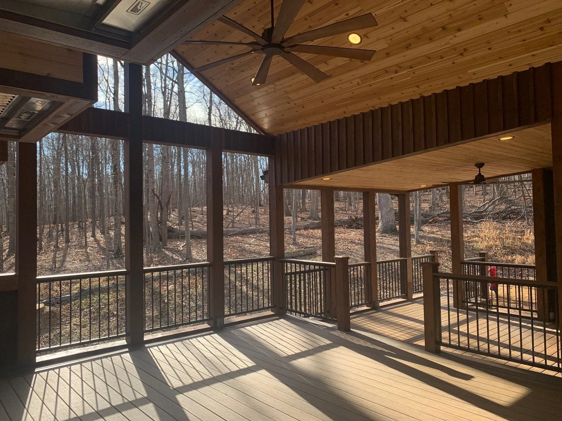 Wooden porch with railing overlooking a wooded area with leafless trees, sunlight casting shadows on the floor, ceiling fans, and recessed lighting.