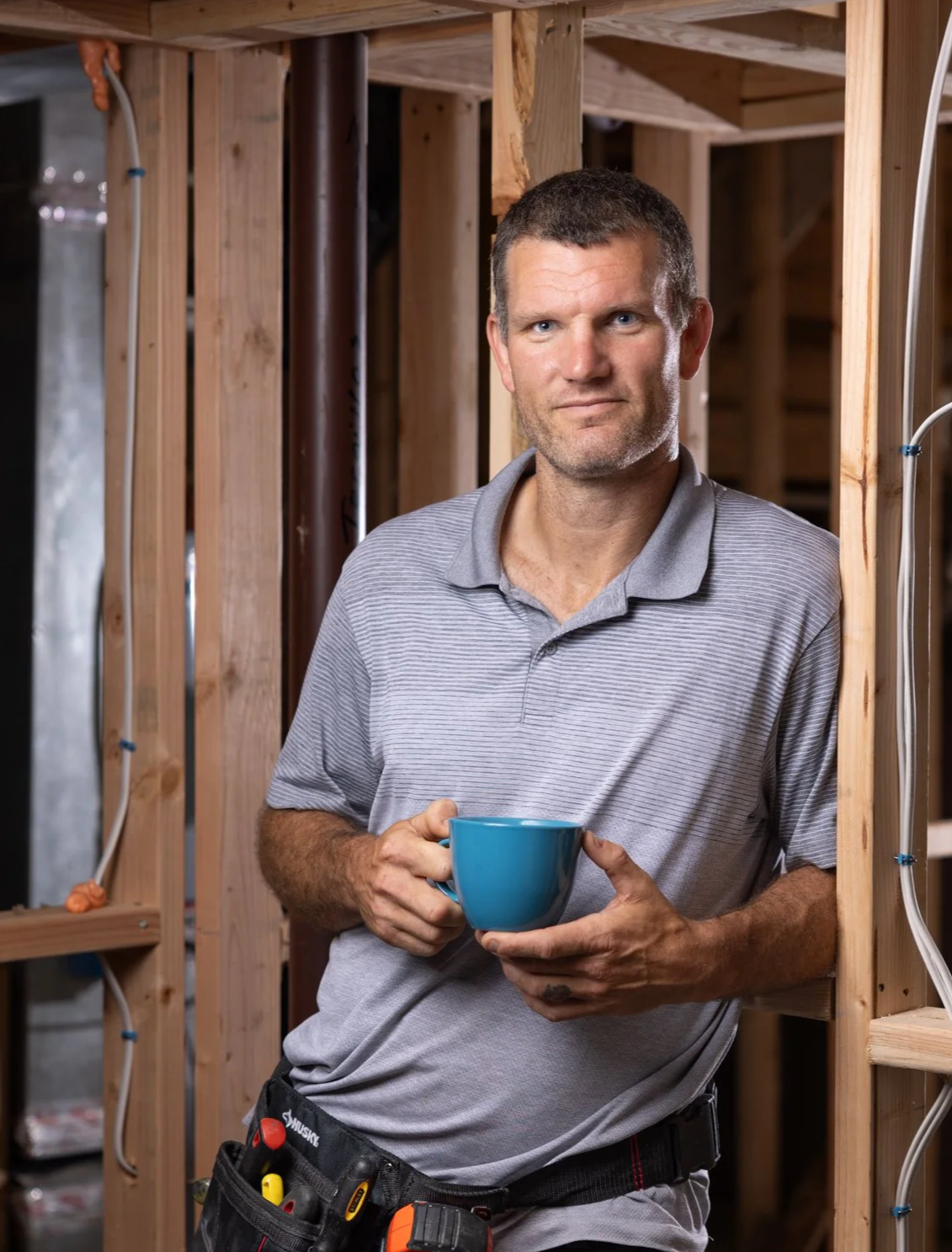 A man in a construction site holding a blue coffee mug, wearing a grey polo shirt and tool belt, standing among unfinished wooden framing.