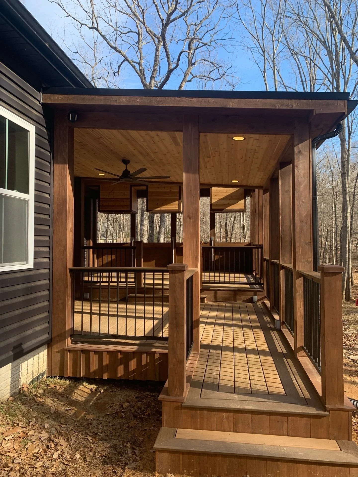 Wooden porch with railings, ceiling fan, and recessed lighting attached to a house surrounded by leafless trees.