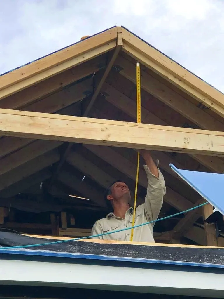 A man working on a building's roof frame, measuring with a yellow measuring tape.