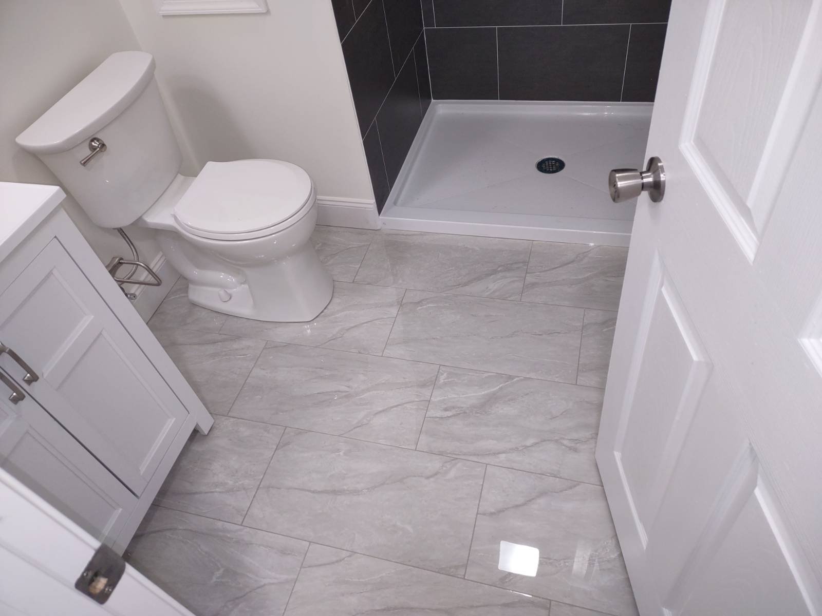 A bathroom with a white toilet, a white vanity, and a shower with a black tiled wall. The floor has light gray marble tiles, and the bathroom door is partially open.