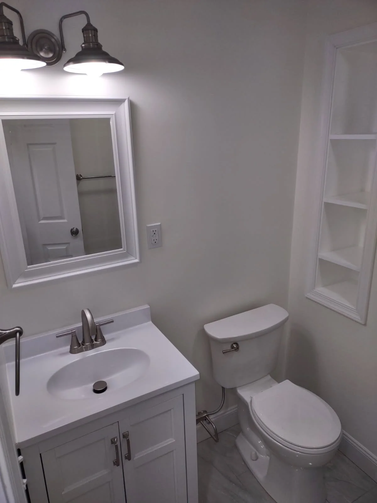 A small bathroom with a white vanity sink, mirror, toilet, and built-in shelves, illuminated by overhead lights.