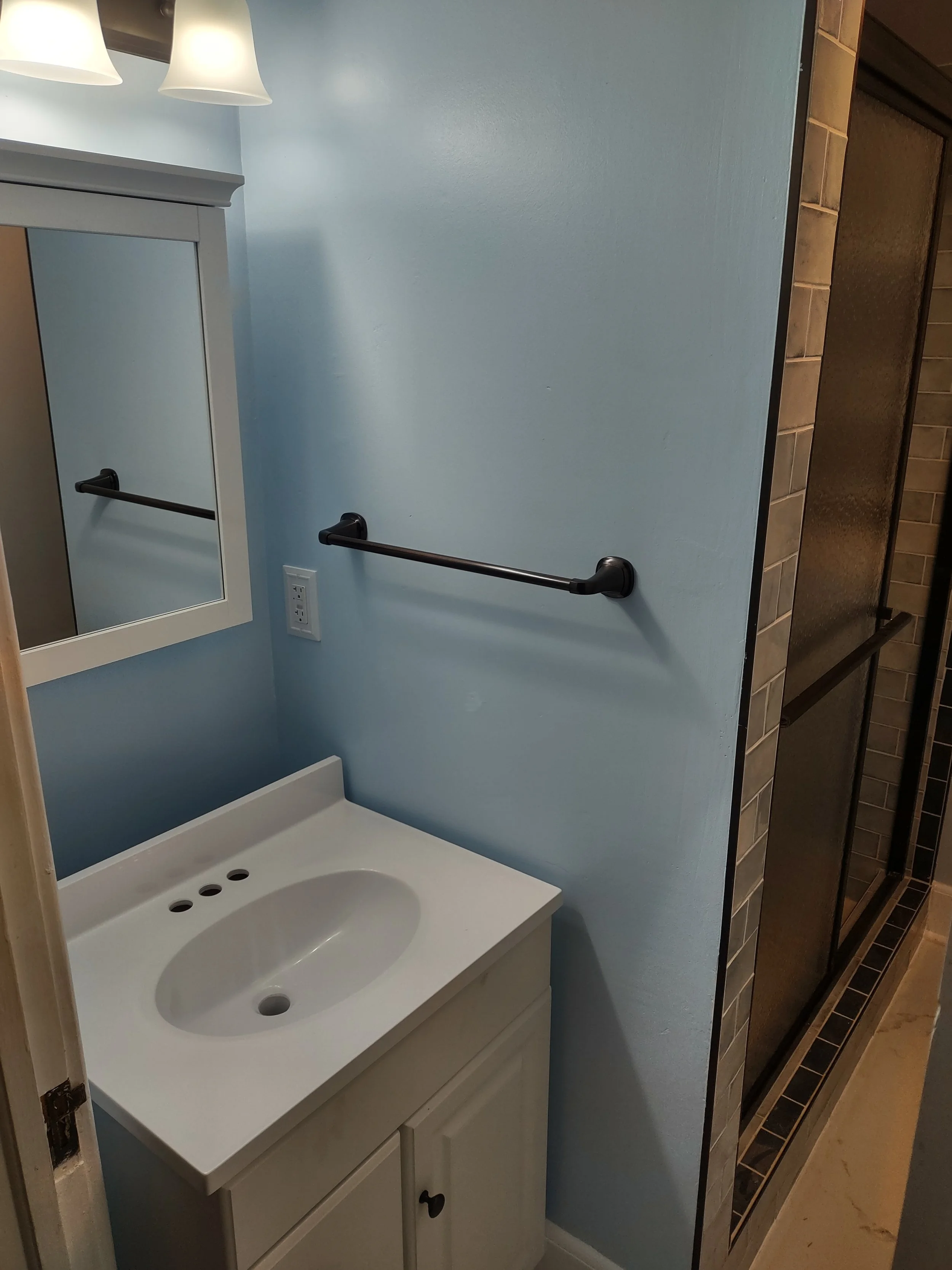 Bathroom with a light blue wall, white vanity with a sink, a mirror cabinet, black towel bar, and a shower with glass door and brown square tiles.