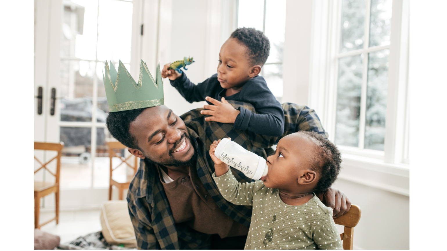 Happy man wearing a green paper crown with a gold glitter stripe, children celebrating indoors with large windows, one child feeding the other from a baby bottle and another child playing with a toy dinosaur.