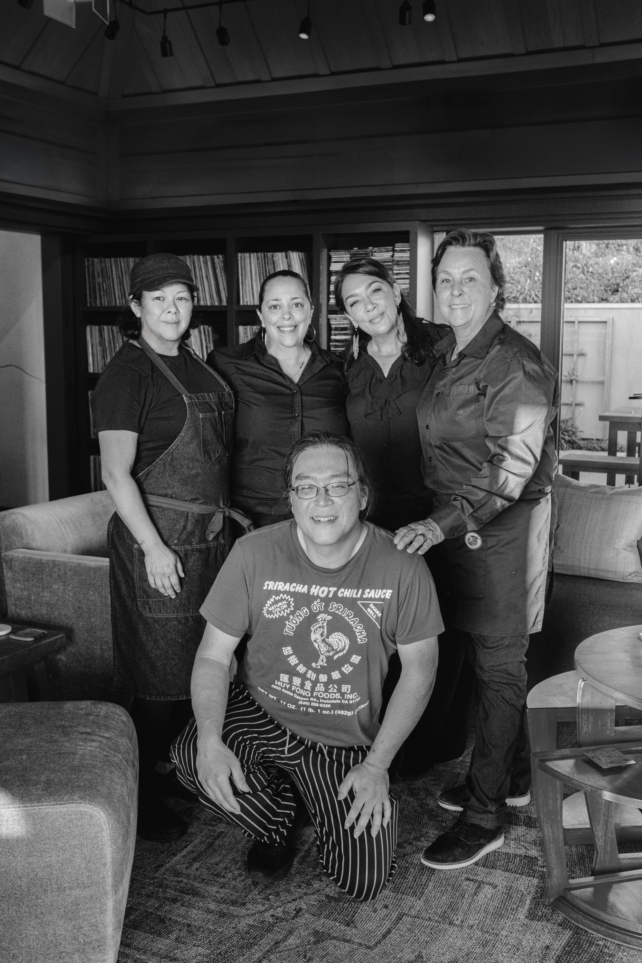 A group of five people, four women and one man, gathered inside a room with wooden shelves and large windows, smiling at the camera.