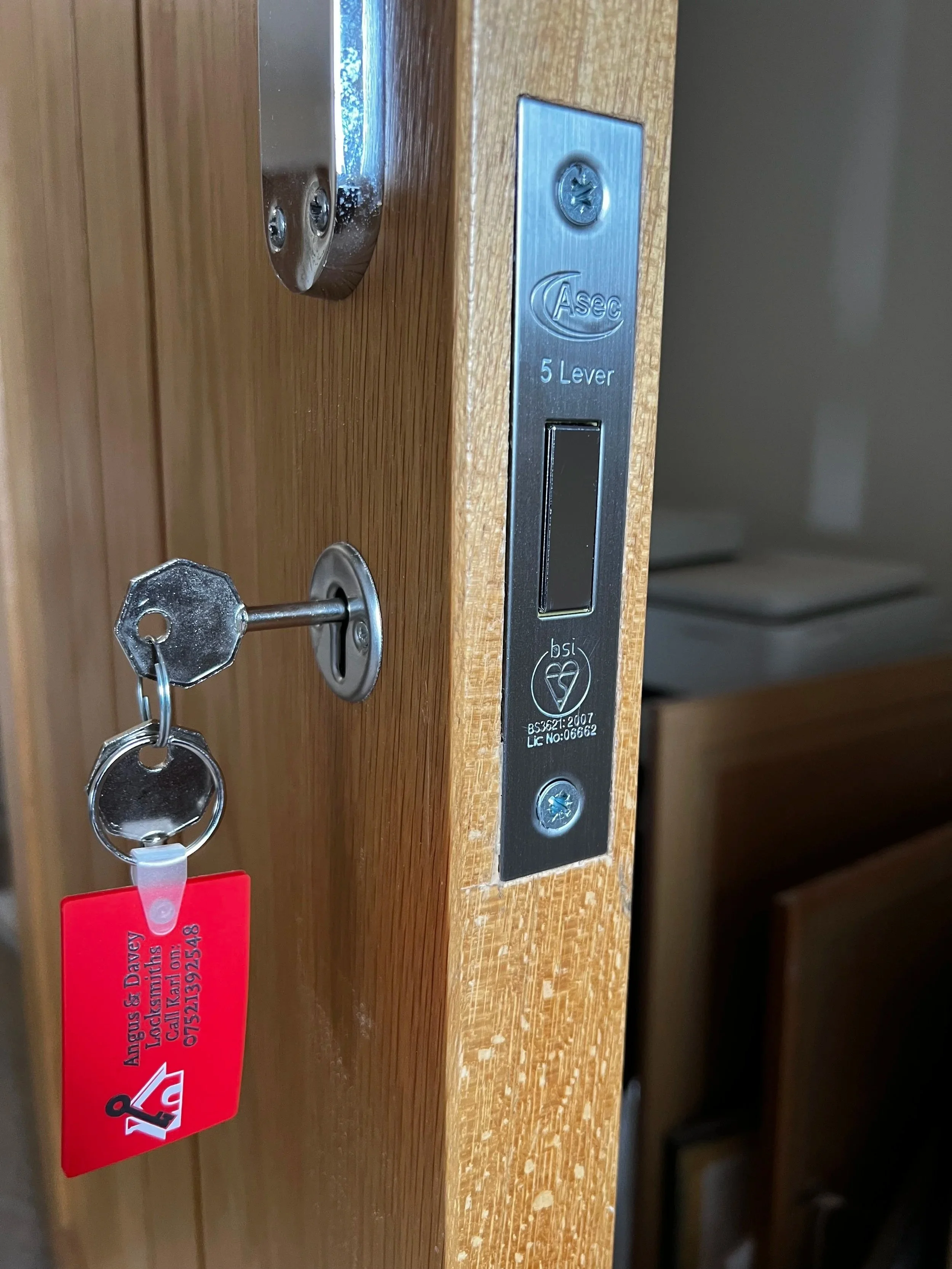 Close-up of a wooden door frame with a door latch, a key hanging on a keyhole, and a metal strike plate labeled 'Asec 5 Lever'.