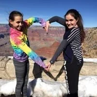 Two young women holding hands and forming a heart shape with their arms near the Grand Canyon, with a dog lying in front of them on a leash.