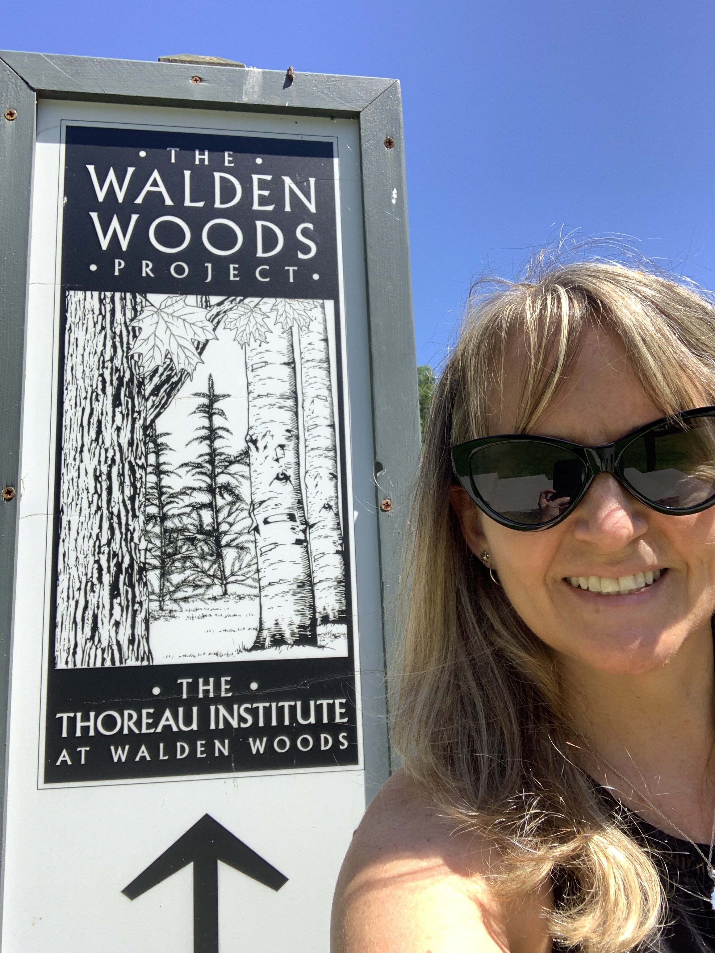 Smiling woman wearing sunglasses taking a selfie next to a sign for Walden Woods and Thoreau Institute at Walden Woods under a clear blue sky.