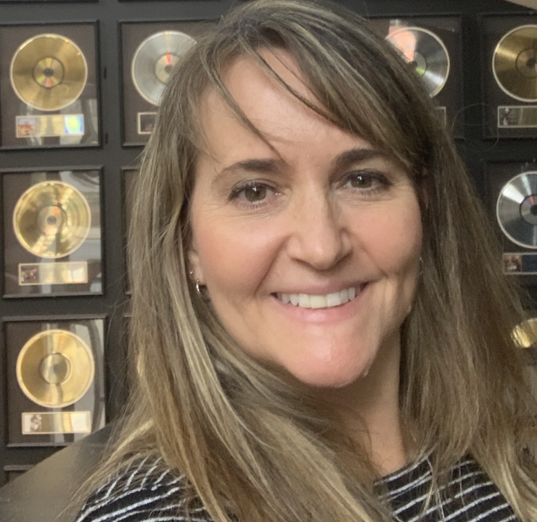Smiling woman with long, light brown hair standing in front of a wall decorated with gold and silver record awards.