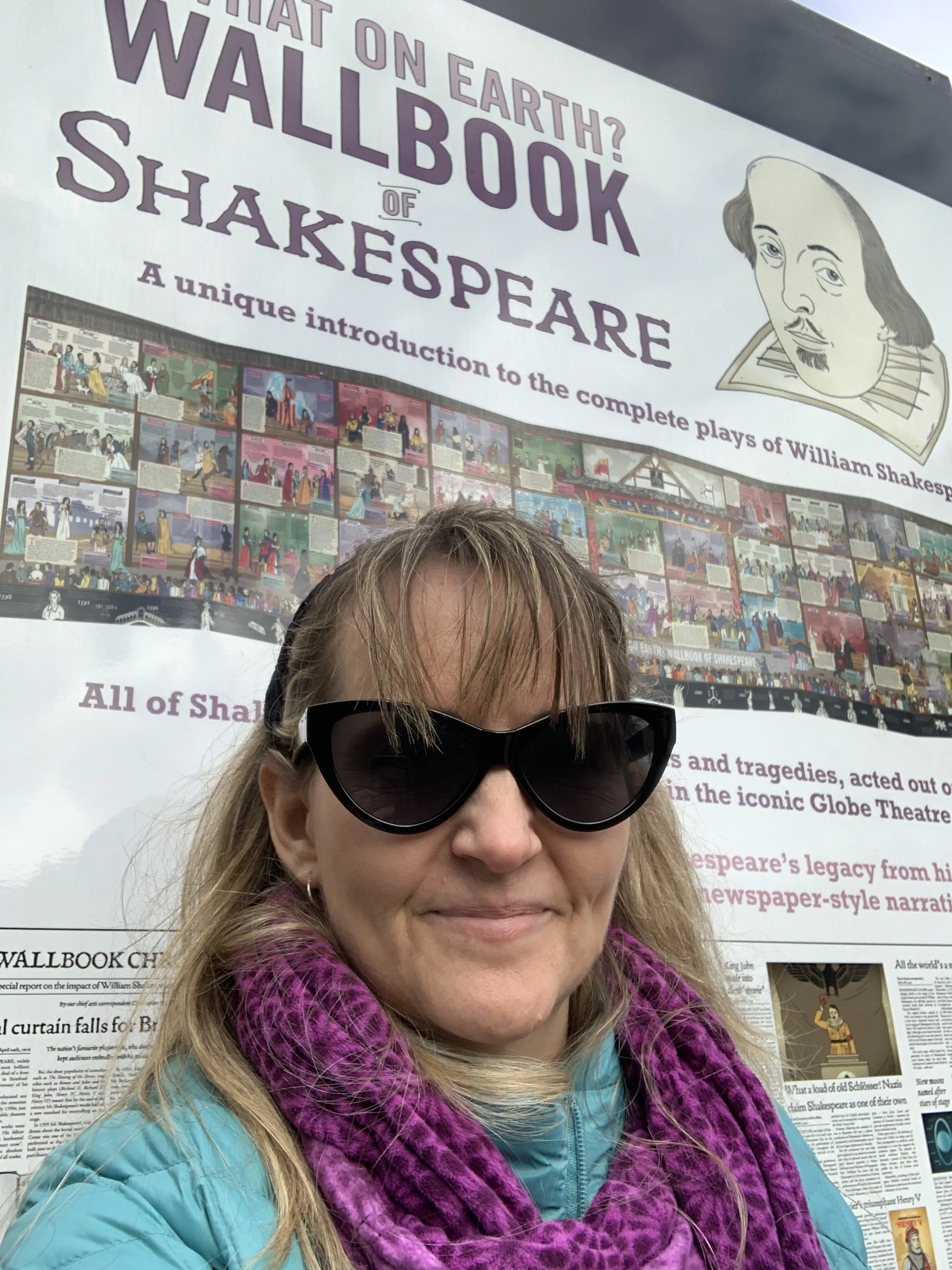 A woman wearing sunglasses, a colorful scarf, and a blue jacket taking a selfie in front of a poster about William Shakespeare's wall book of plays.